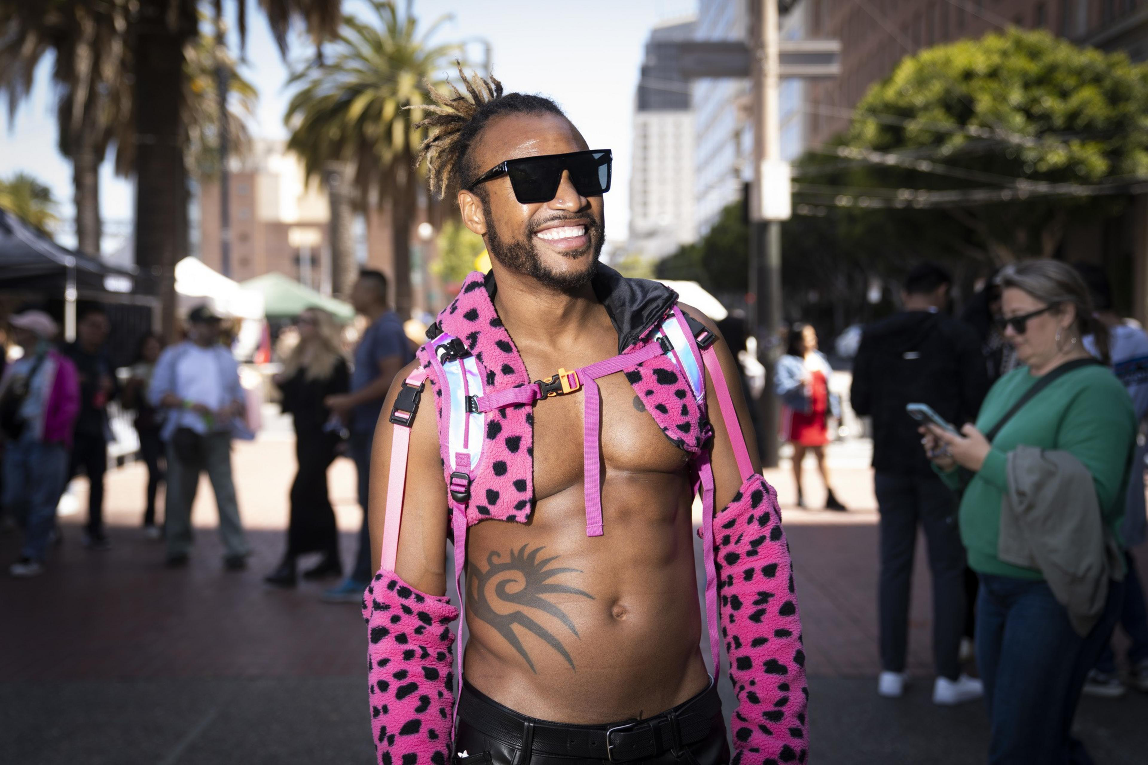 A smiling person in sunglasses wears a pink leopard-print vest and sleeves, displaying a tribal tattoo on their chest. They stand in a busy, sunlit urban setting.