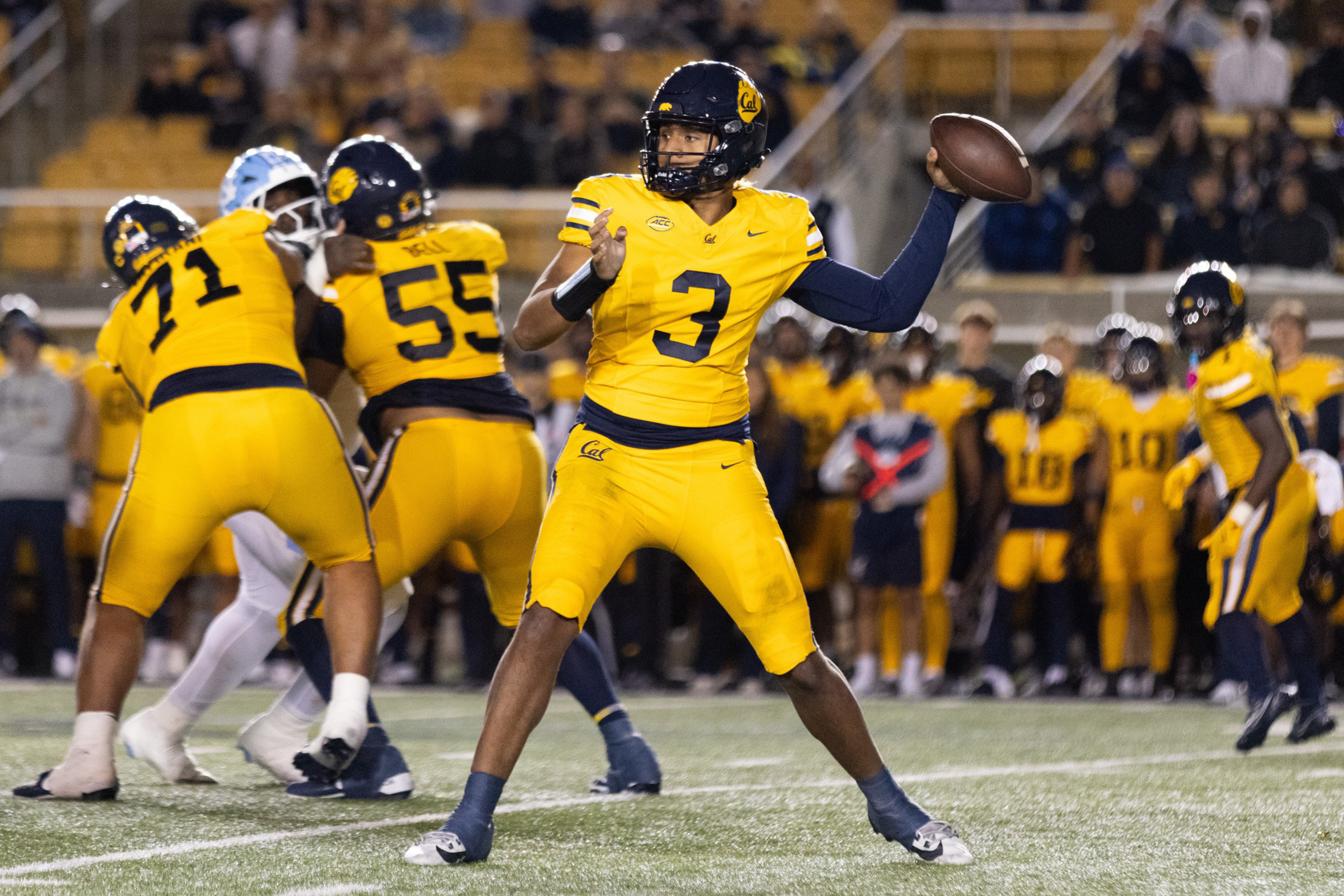 A football player wearing a yellow uniform with number 3 prepares to throw a pass while teammates block defenders on the field.