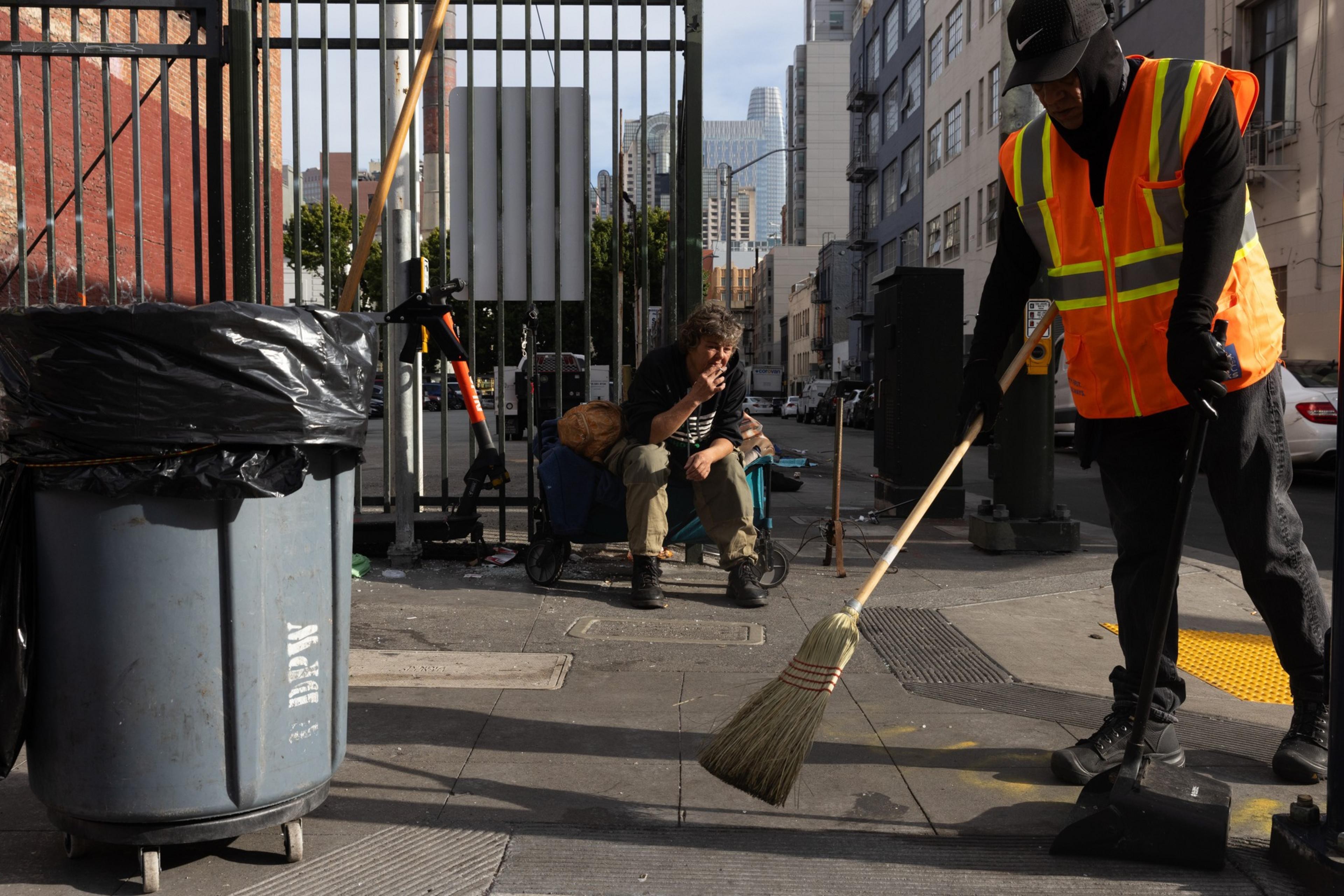 A homeless woman sits on the street beside a city worker. 