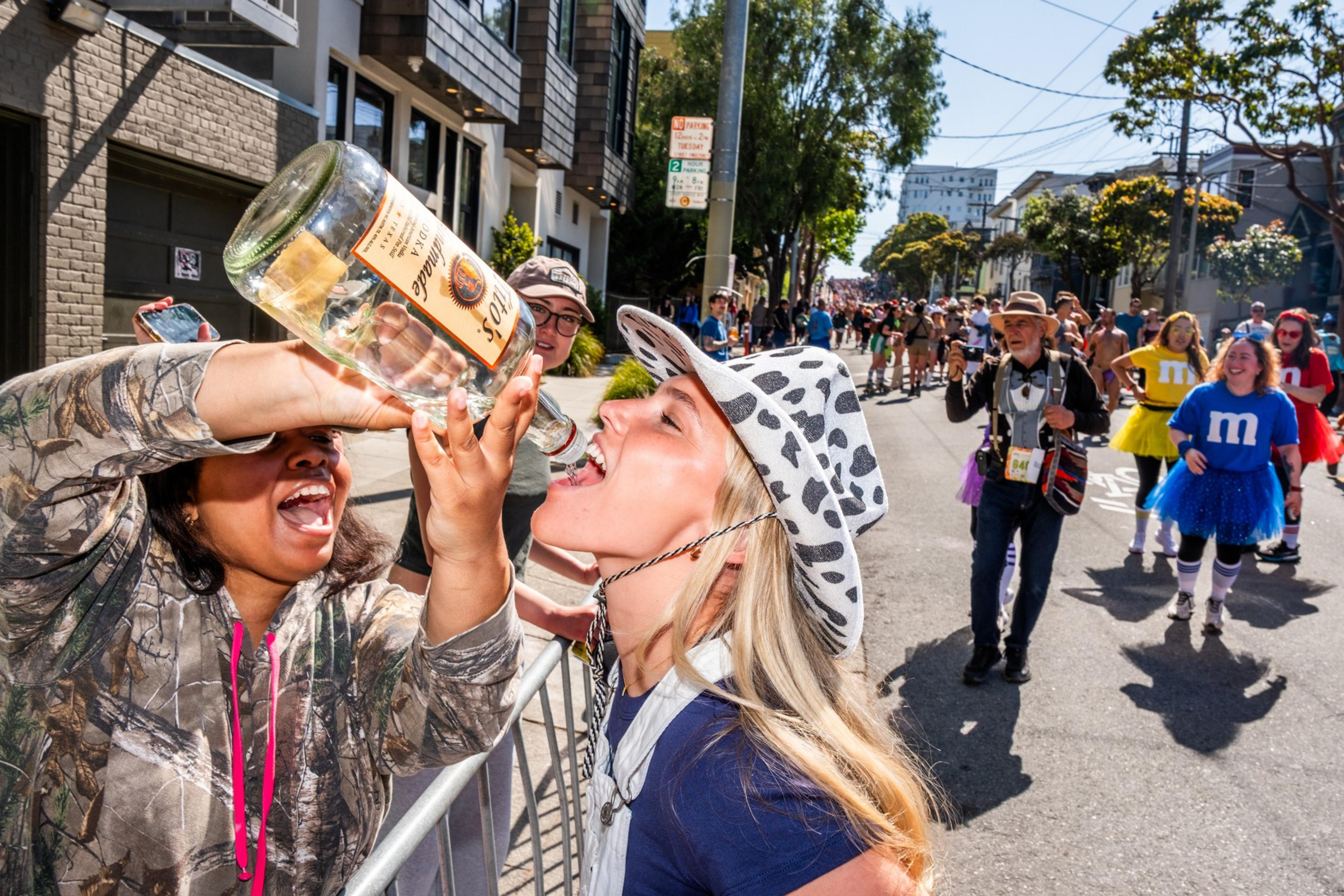 A person in a camo hoodie pours from a large bottle into the mouth of a woman in a spotted hat. In the background, people in colorful costumes walk down the street.