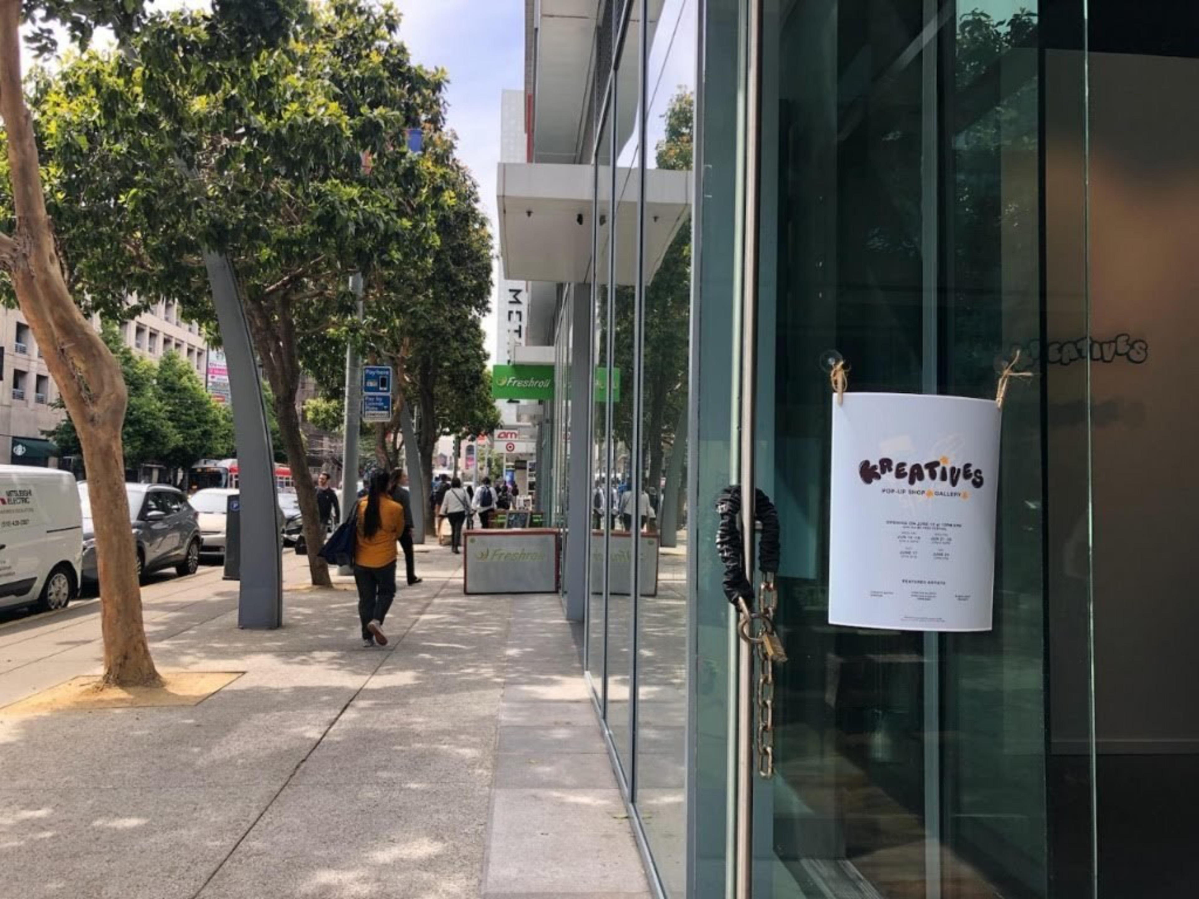 A city sidewalk with pedestrians, trees, parked cars, and a glass storefront with a "KREATIVES" pop-up shop sign on the door.