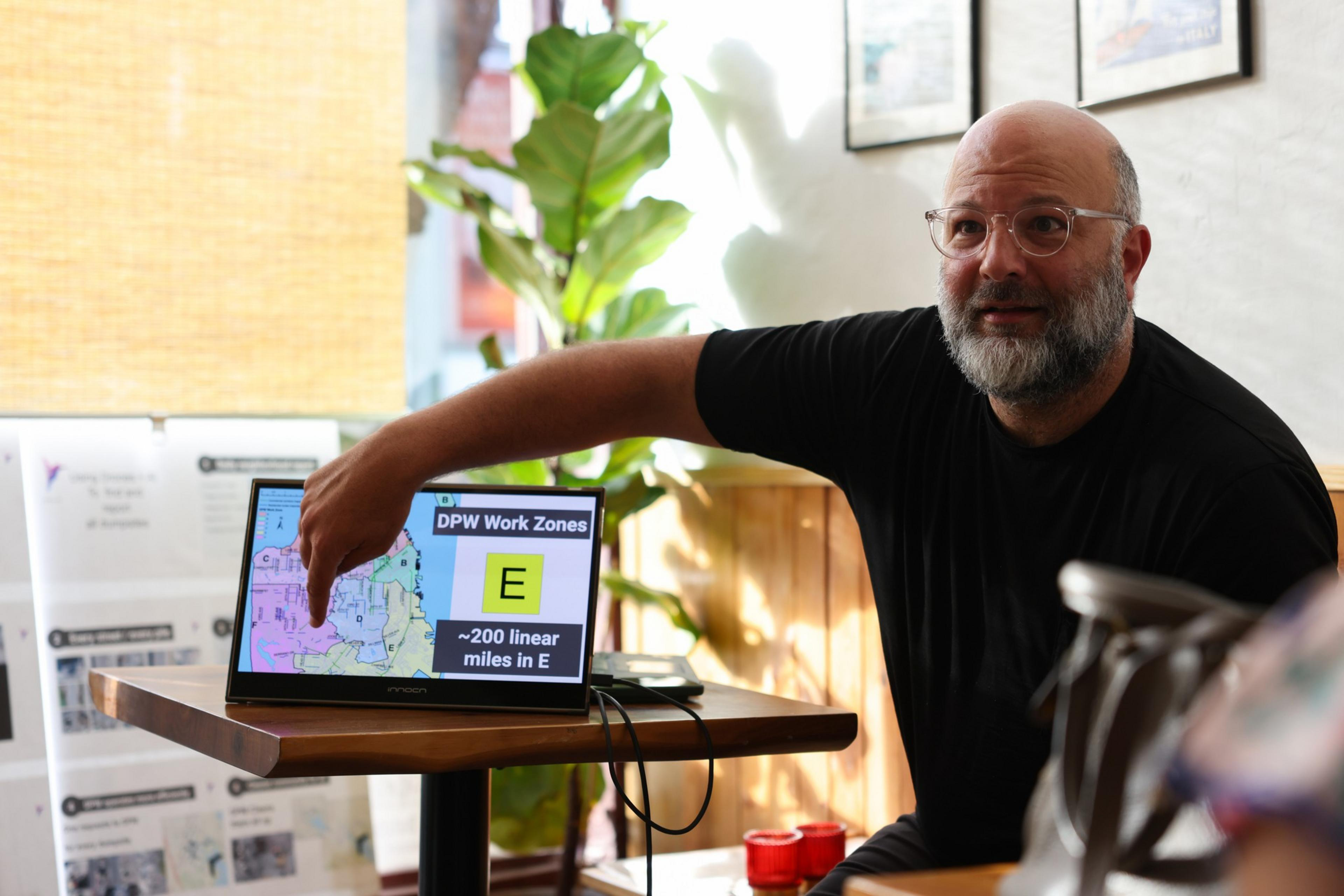 A bearded man in glasses points at a laptop screen displaying a map labeled "DPW Work Zones E" with "~200 linear miles in E" on it.