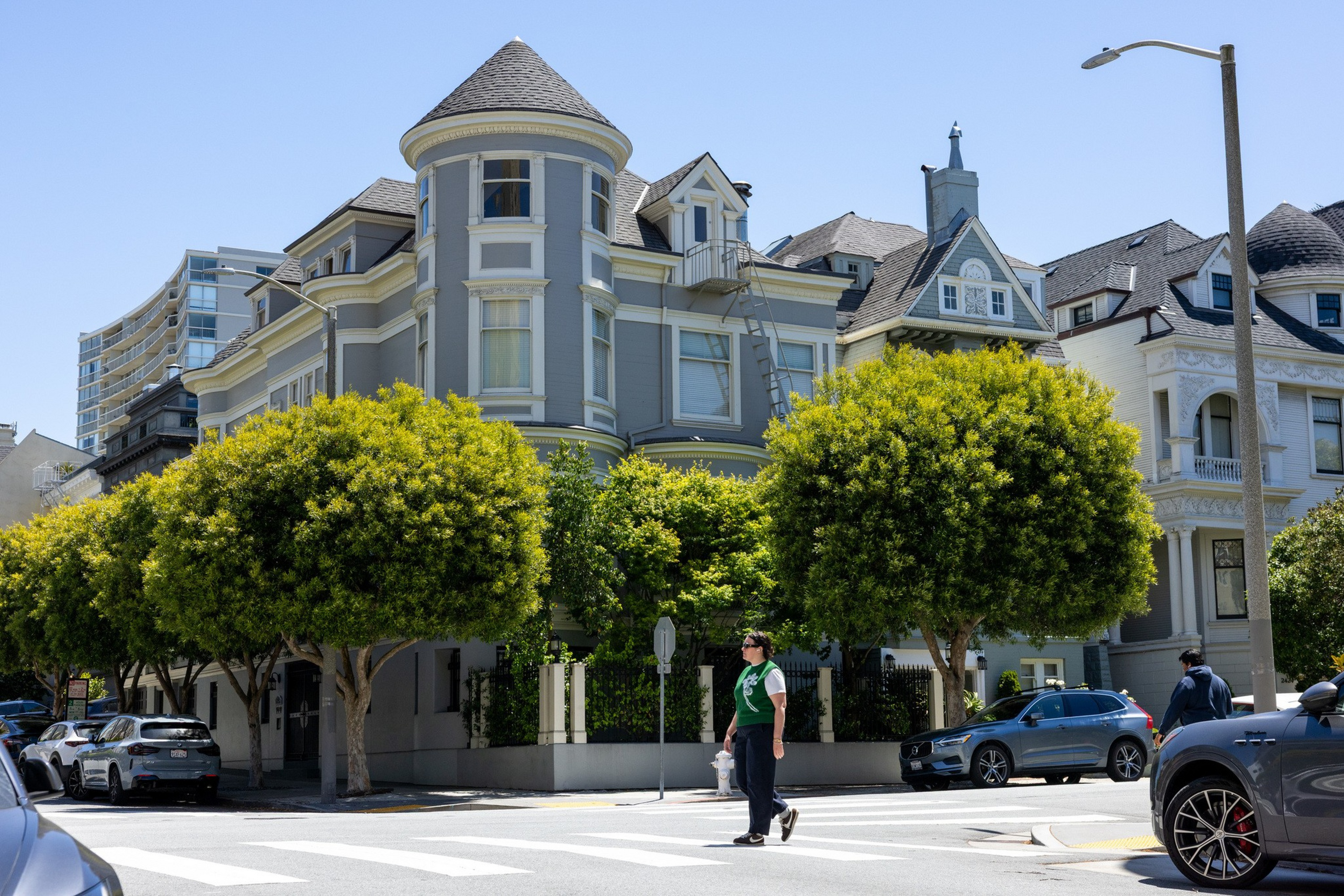 A person wearing a green shirt crosses a street in front of a large, gray Victorian-style house with a rounded turret and green trees lining the sidewalk.