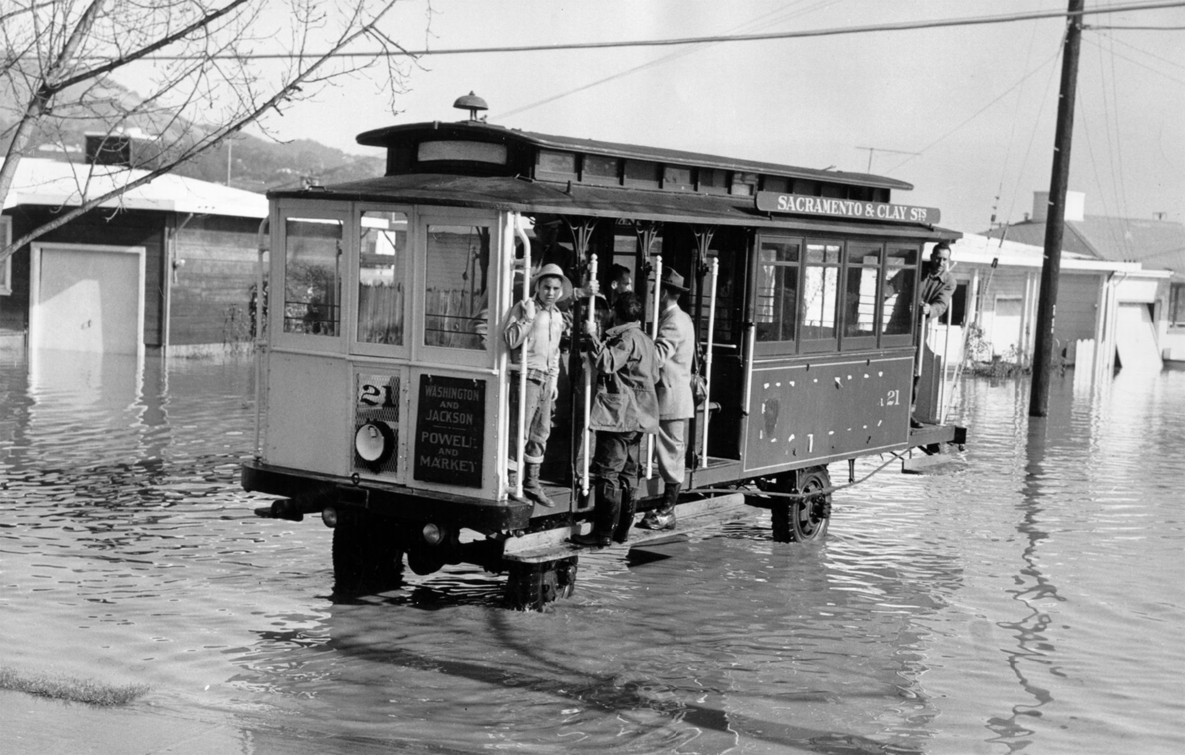 A streetcar with passengers stands in flooded water, with water reaching above the wheels and people hanging onto the sides.