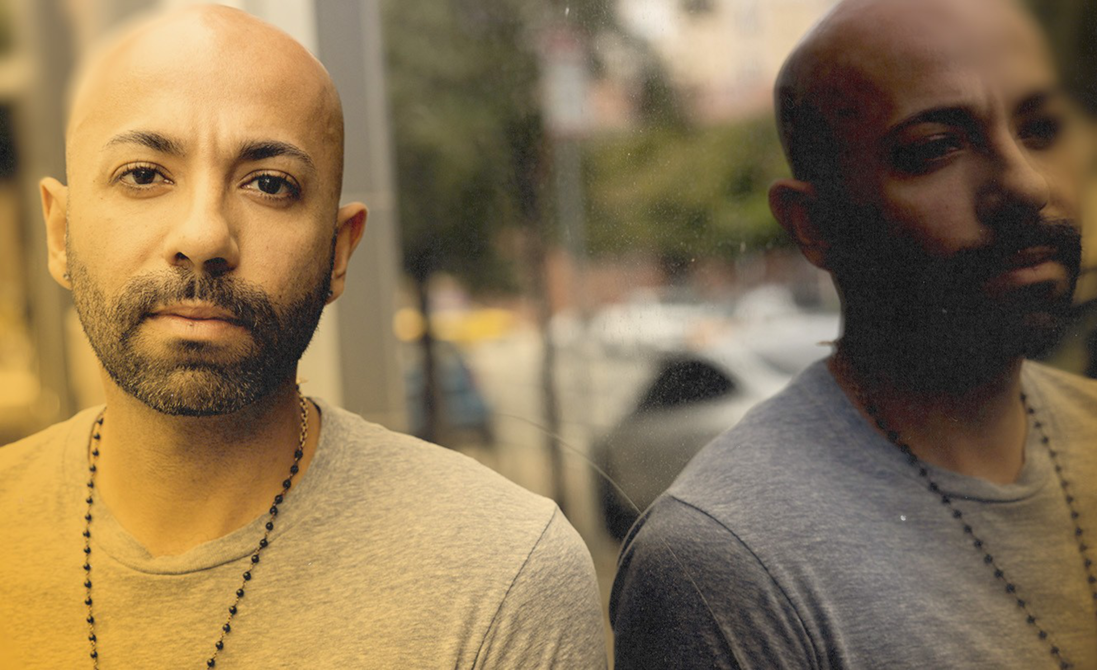 A bald man with a beard and a beaded necklace stands close to a reflective glass, showing his serious expression and reflection.