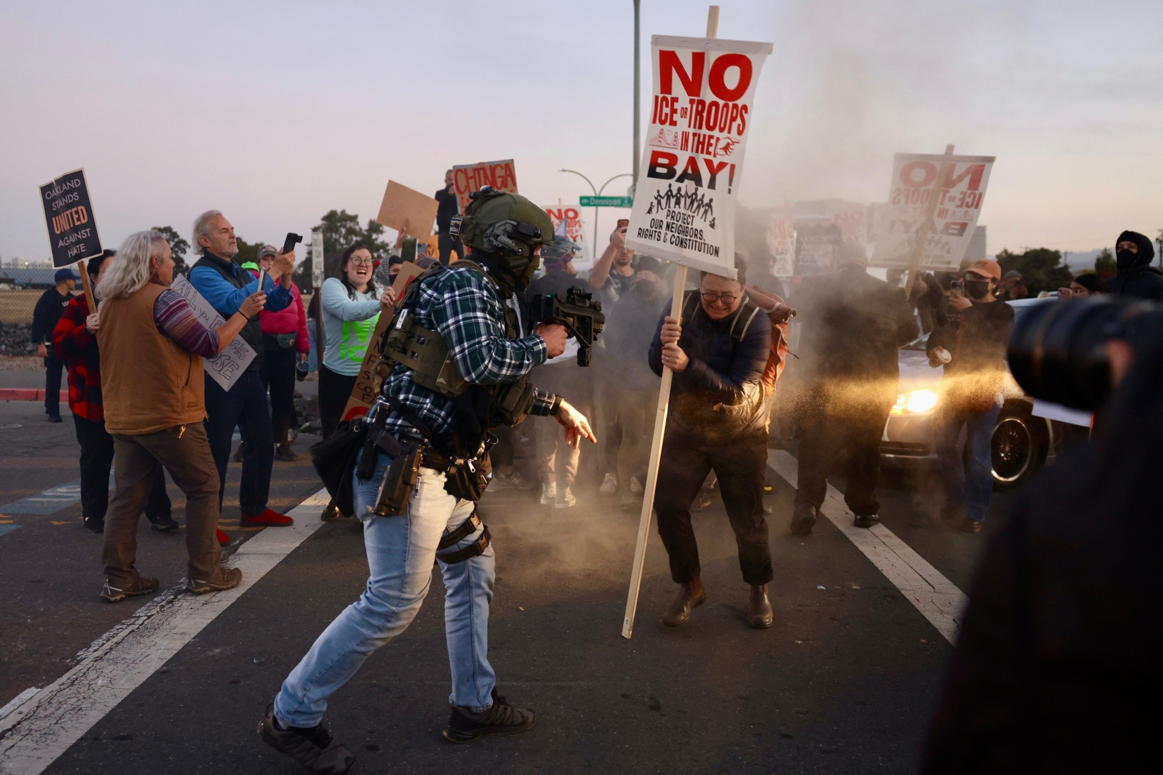 A heavily armed officer in tactical gear fires tear gas at protesters holding signs against ICE troops in the Bay during a tense street confrontation.