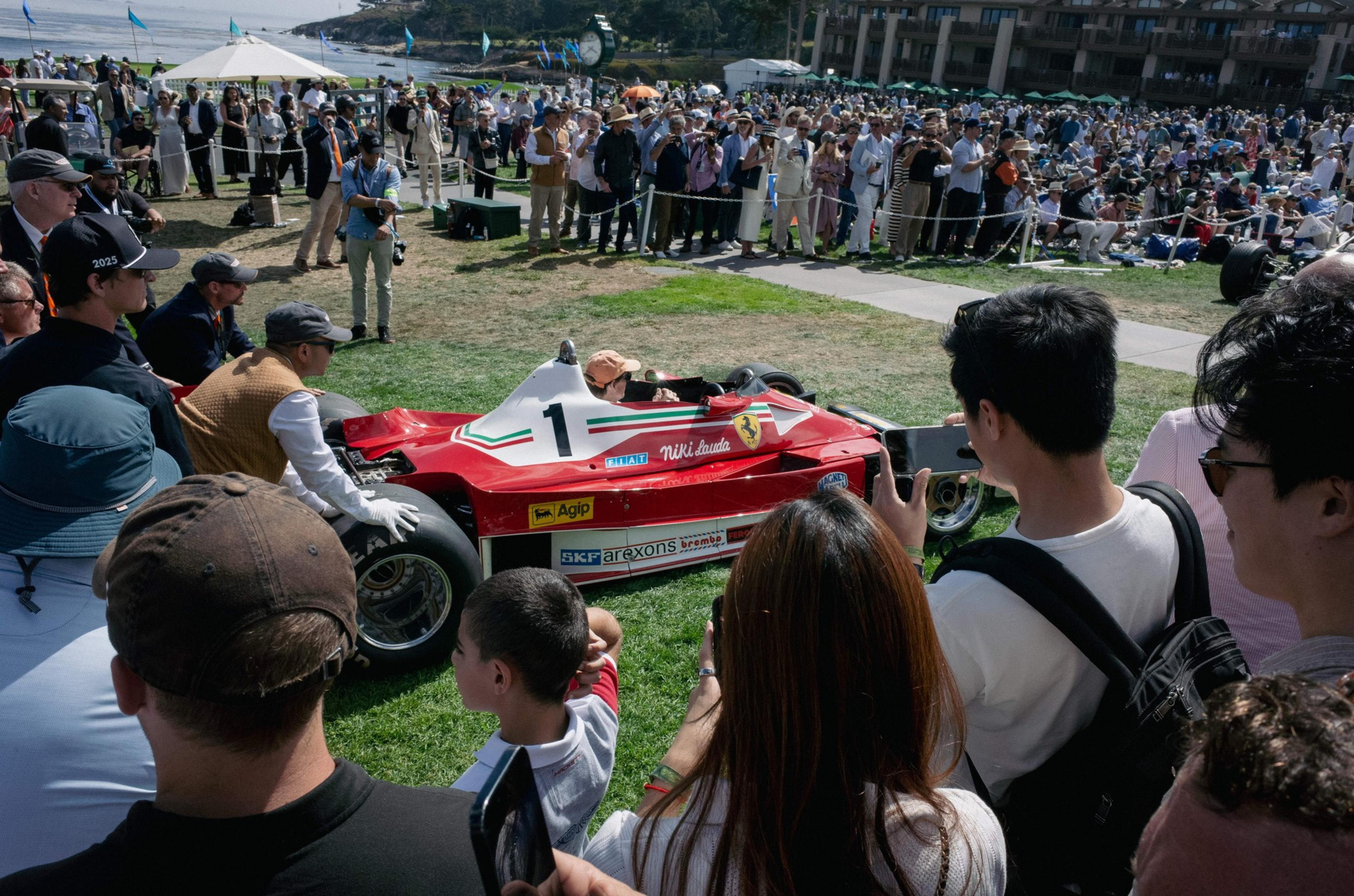 A vintage red Ferrari Formula 1 car with the number 1 and "Niki Lauda" on the side is surrounded by a crowd at an outdoor event near the coast.