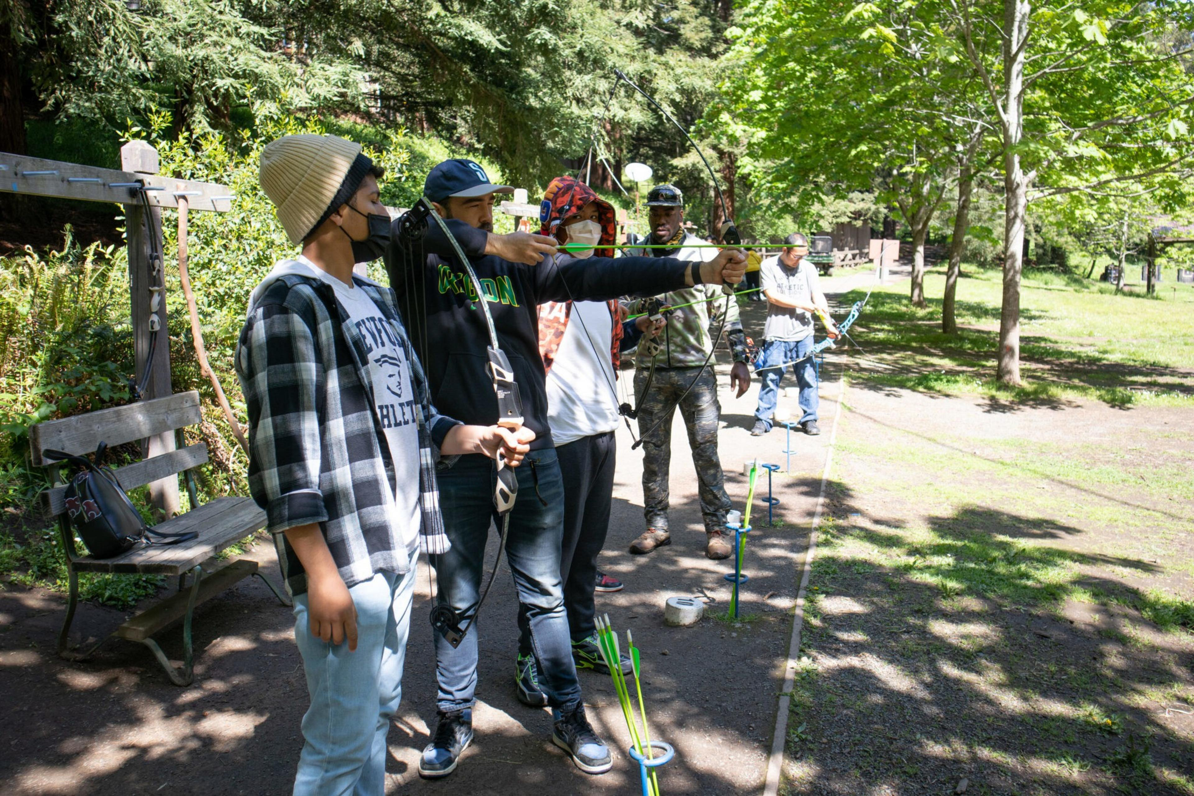 Five people stand in a row outdoors, with three of them aiming bows and arrows, surrounded by green trees and grassy areas.