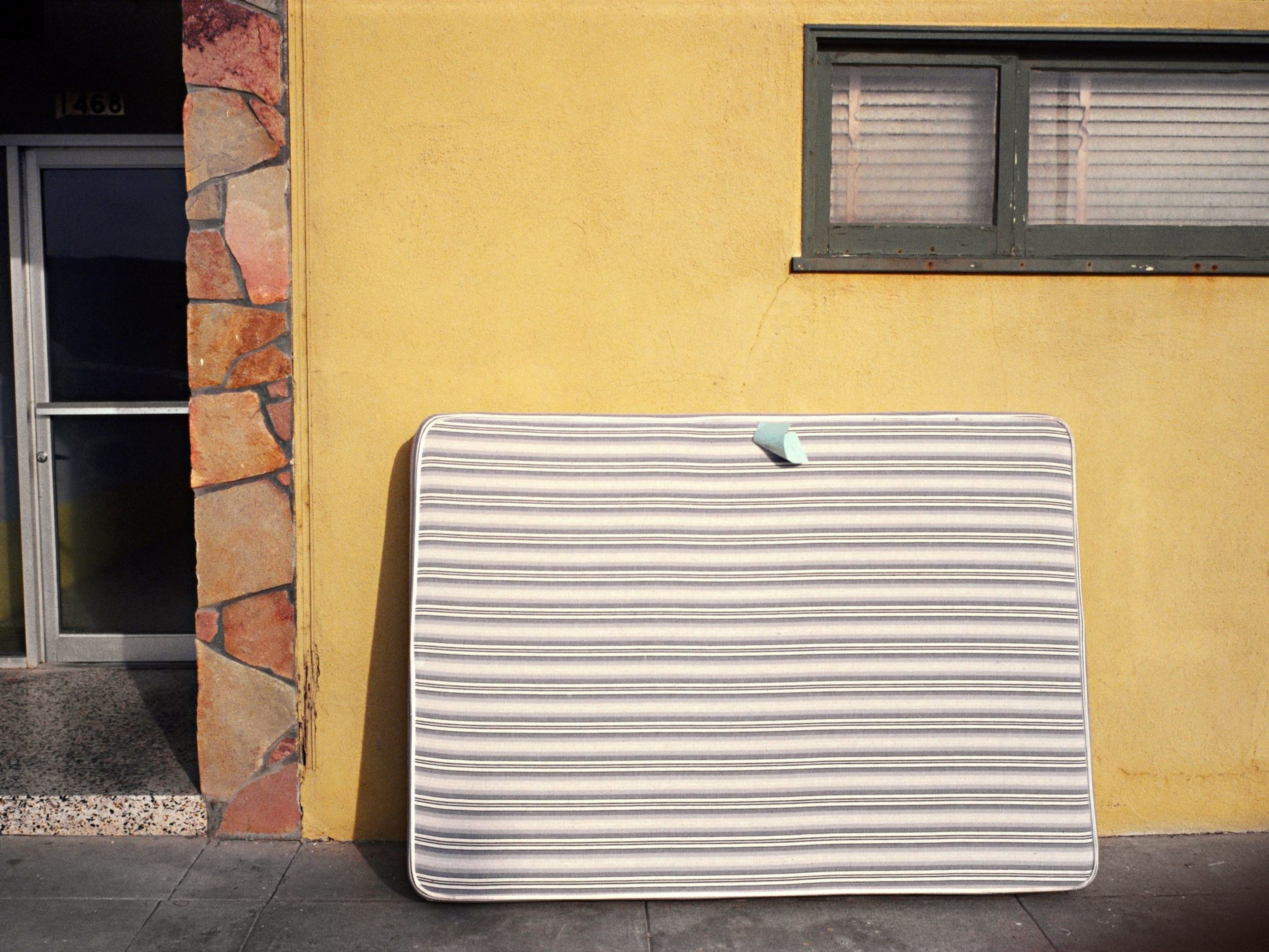 A striped mattress leans against a yellow wall outdoors. Nearby, there's a stone-trimmed glass door. A small window with closed blinds is above the mattress.