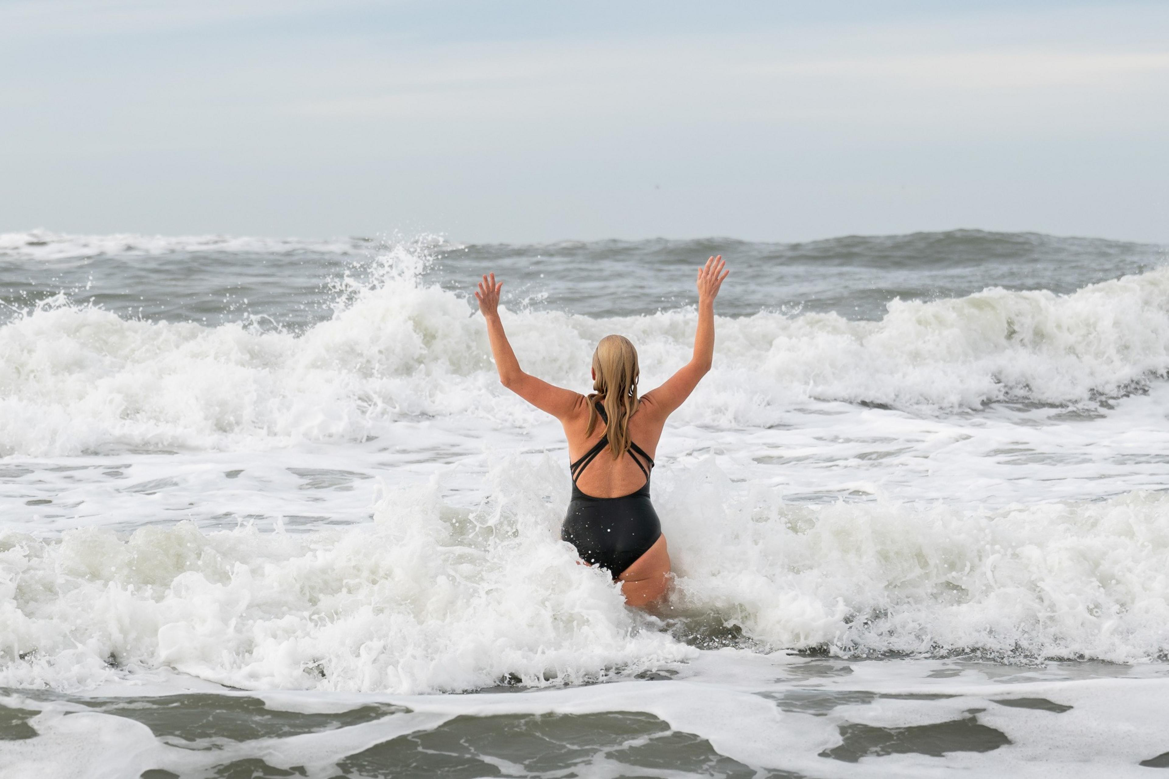 A person in a black swimsuit stands with arms raised, facing crashing ocean waves under a cloudy sky.