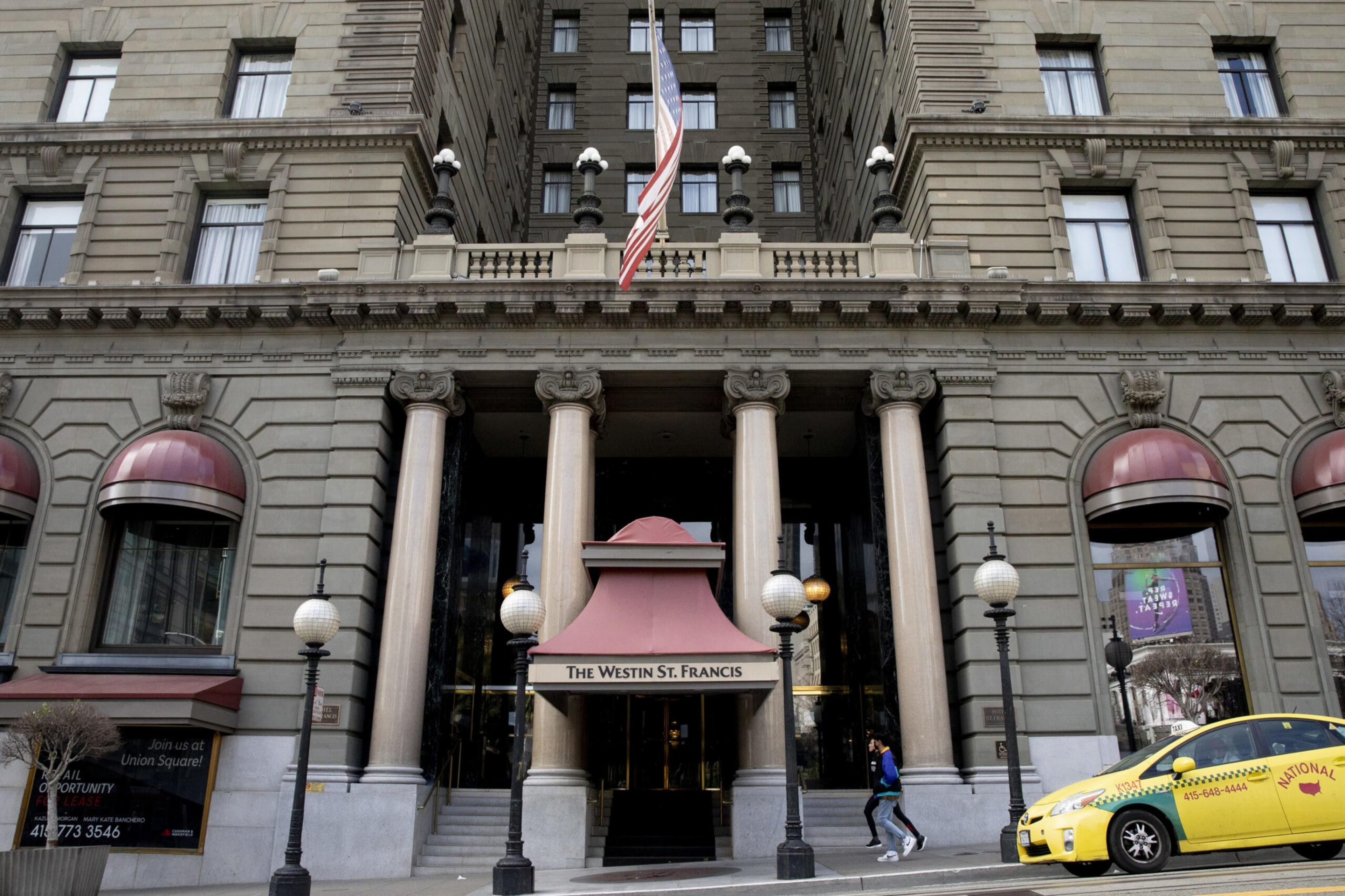 A grand stone building with tall columns and an American flag above the entrance labeled "The Westin St. Francis." A yellow taxi and a pedestrian are nearby.