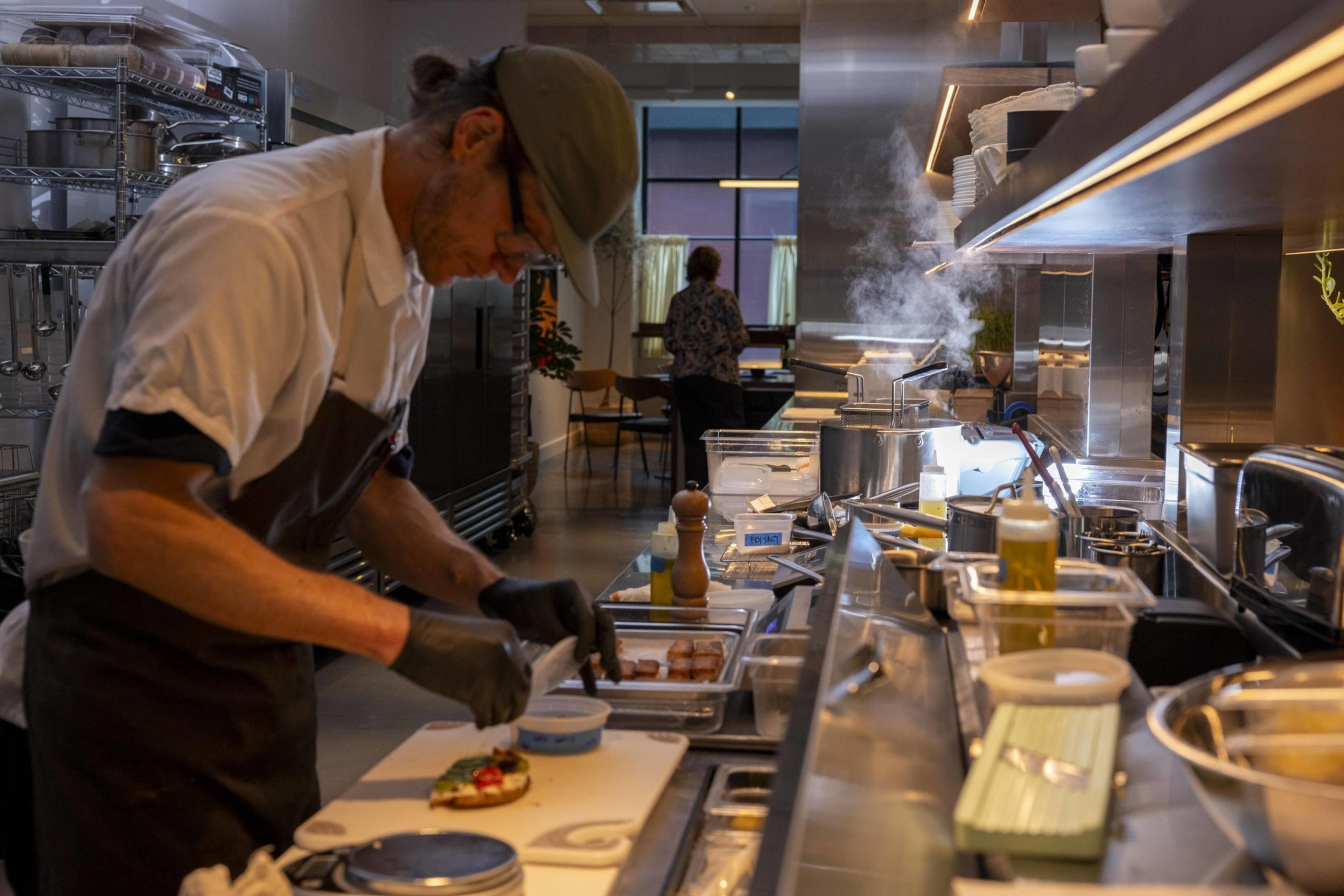 A chef in a kitchen carefully prepares food on a counter. Steam rises from pots nearby, and various cooking utensils and ingredients are scattered around.