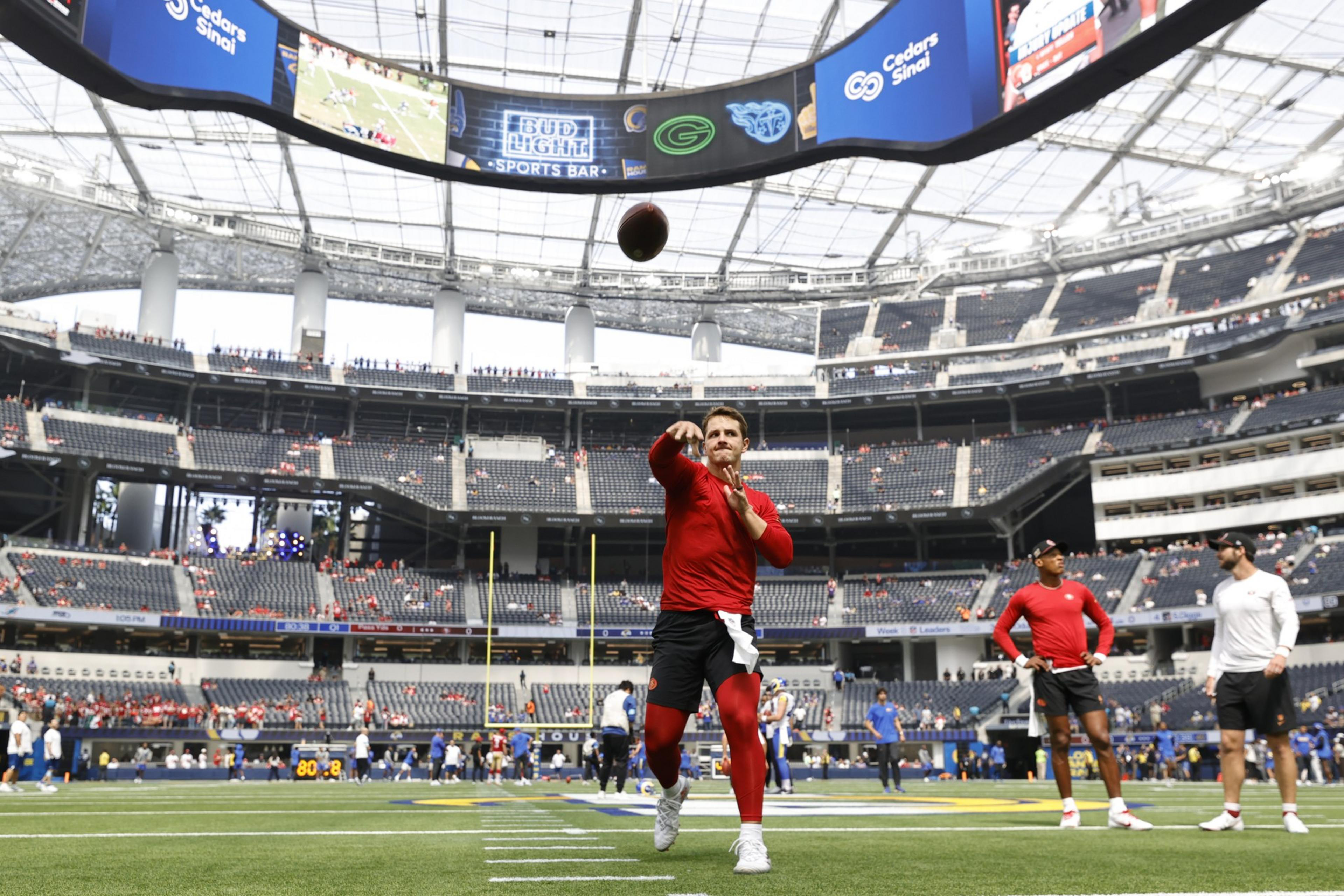 A football player in red practice gear throws a football on a stadium field, with a giant scoreboard above and sparse spectators in the stands.