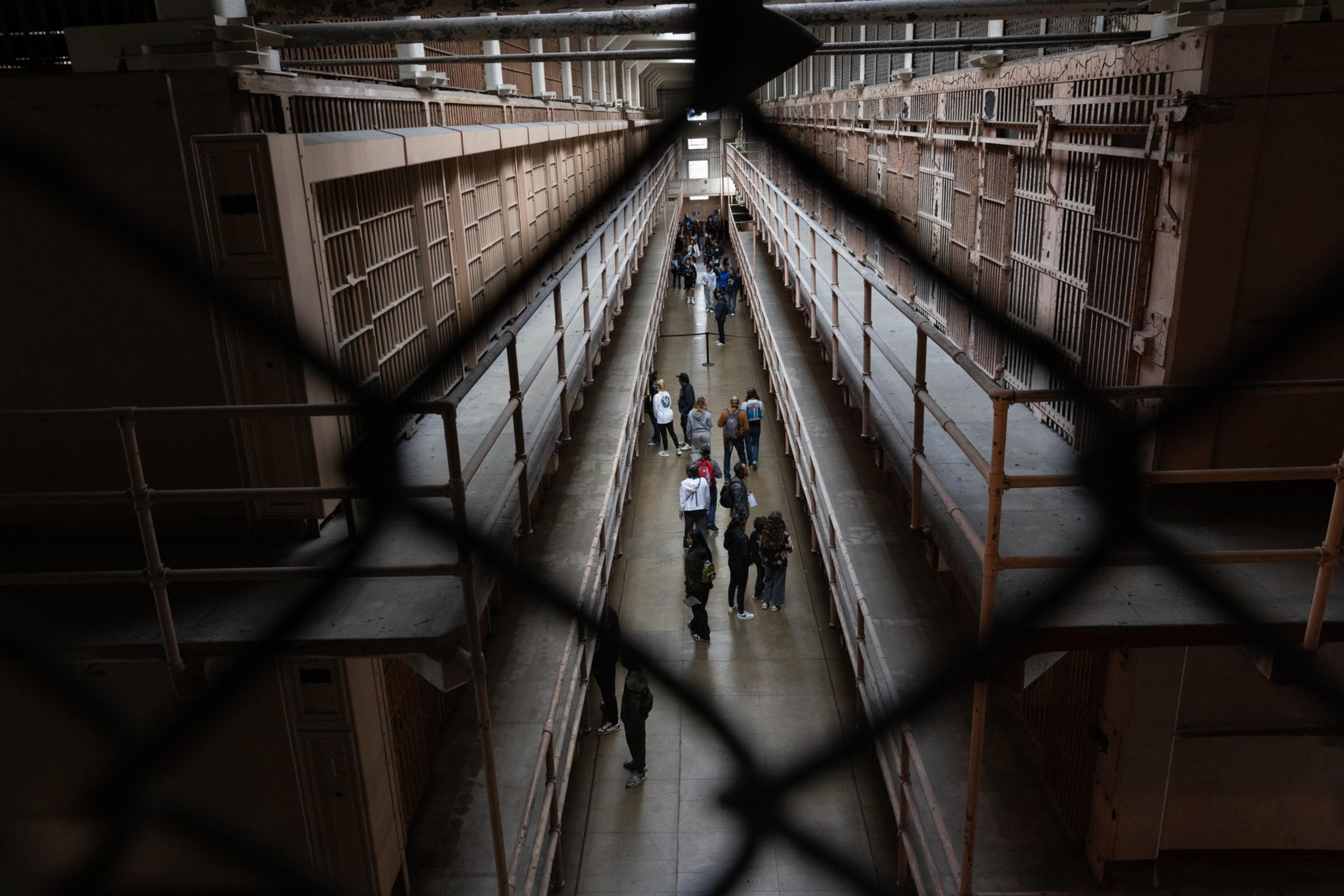 The image shows a prison corridor with barred cells on both sides. A group of people is walking down the central walkway, viewed through a crosshatched fence.