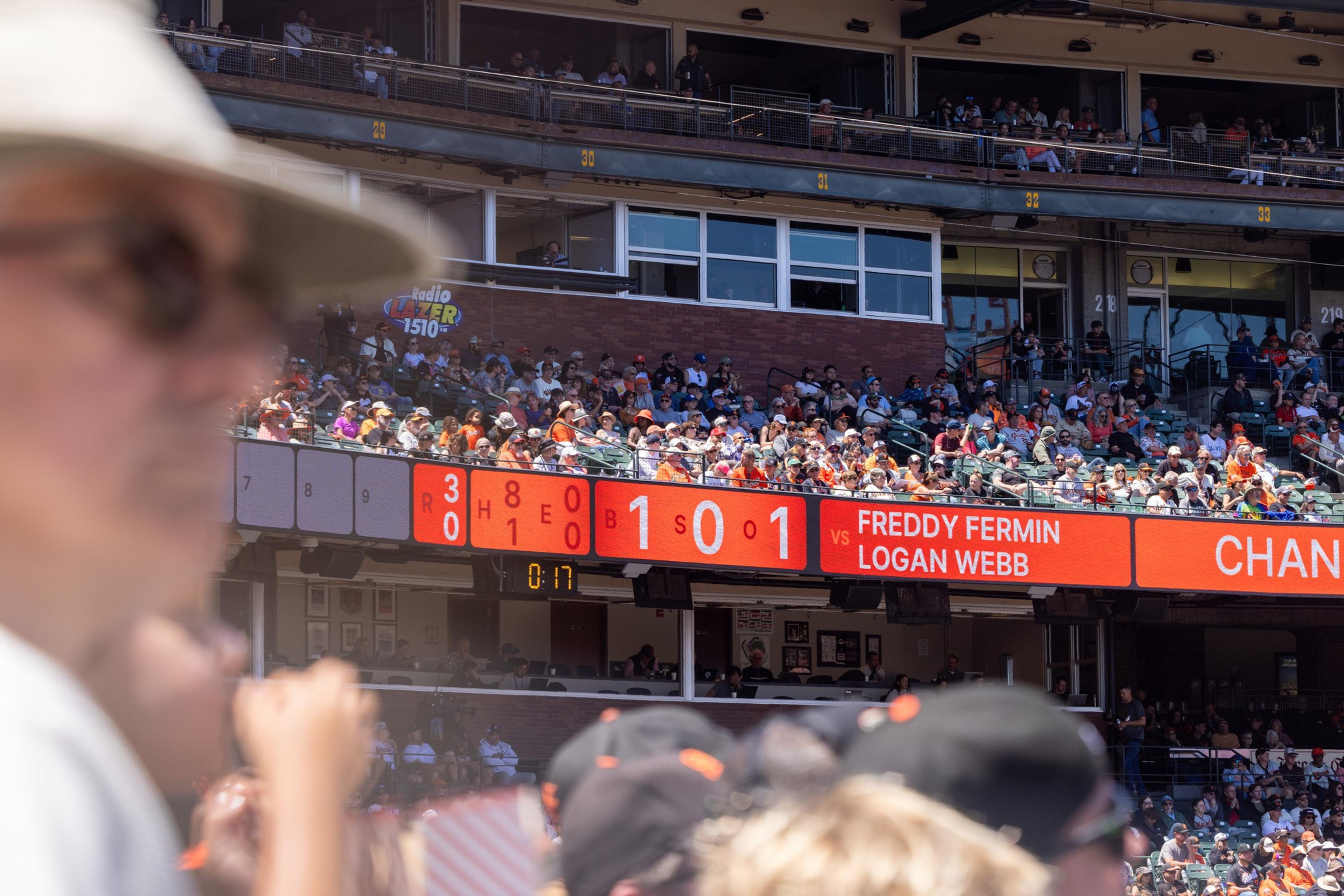 A baseball stadium with a scoreboard displaying game stats and player names, Freddy Fermin vs. Logan Webb. A crowd is seated, and a blurred person is in the foreground.