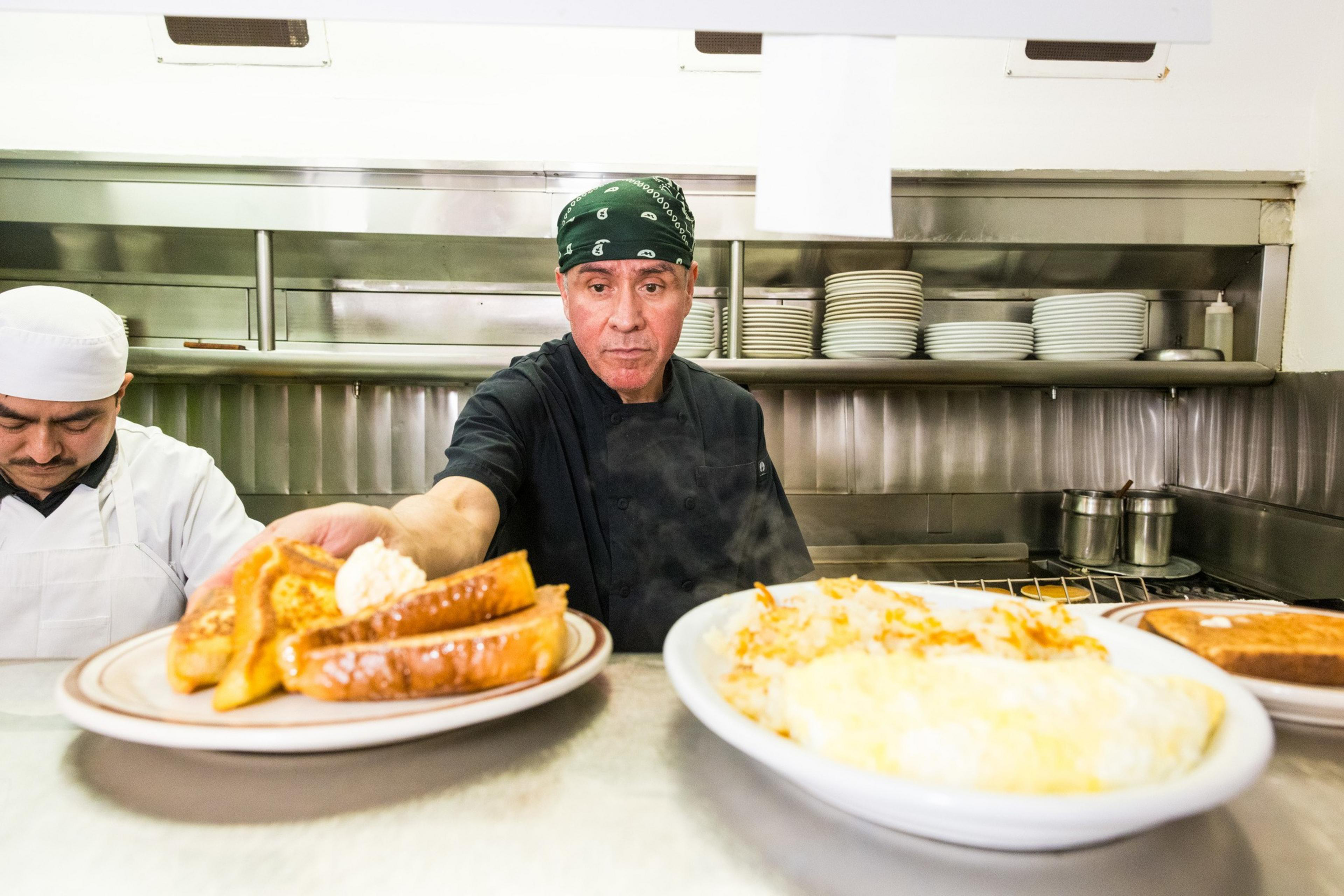 A chef in a kitchen serves a plate of French toast with butter. Another chef works beside him. Plates and cooking pots are visible in the background.