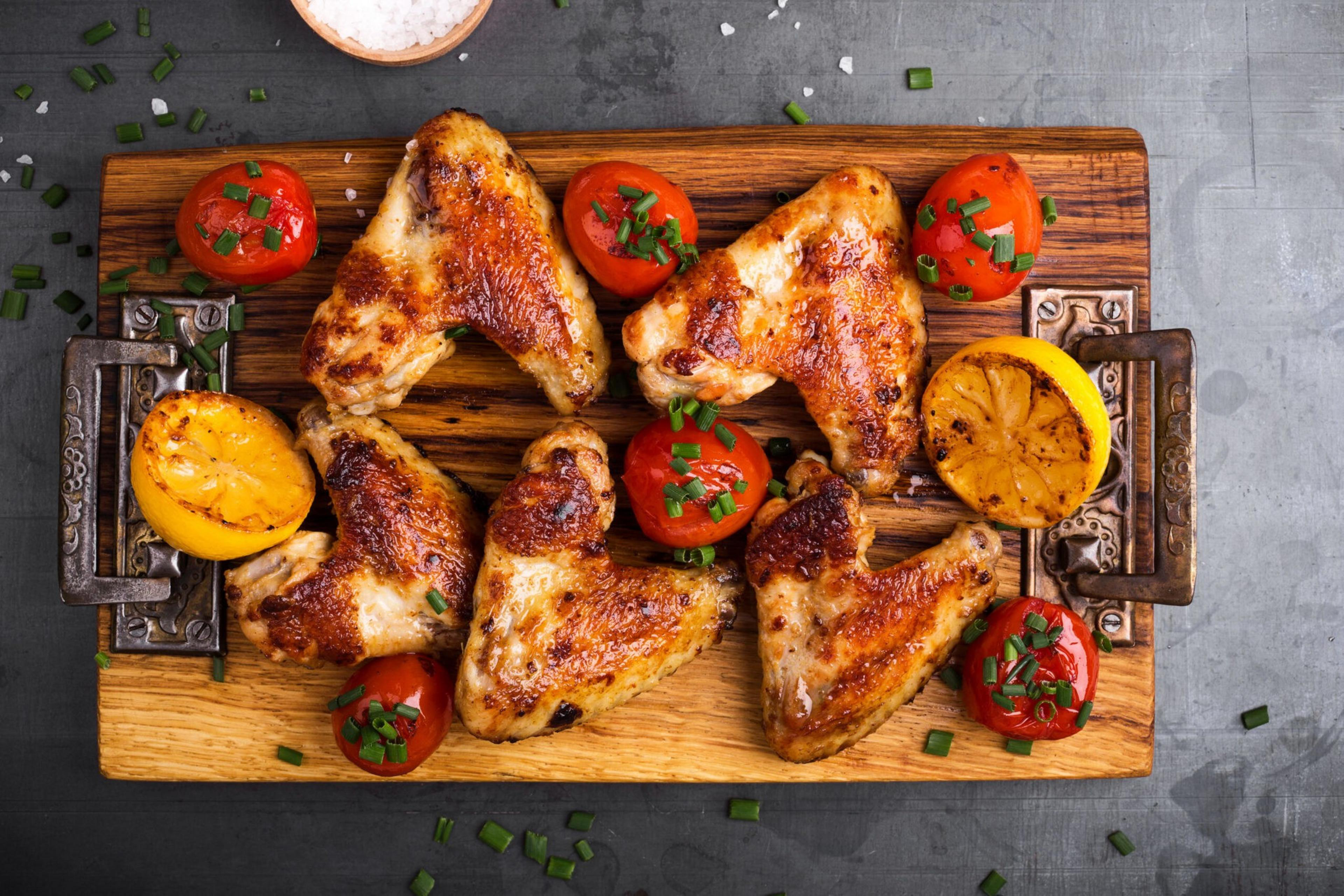 Six grilled chicken wings and six grilled cherry tomatoes garnished with chopped green onions are arranged on a wooden serving board with two grilled lemon halves.