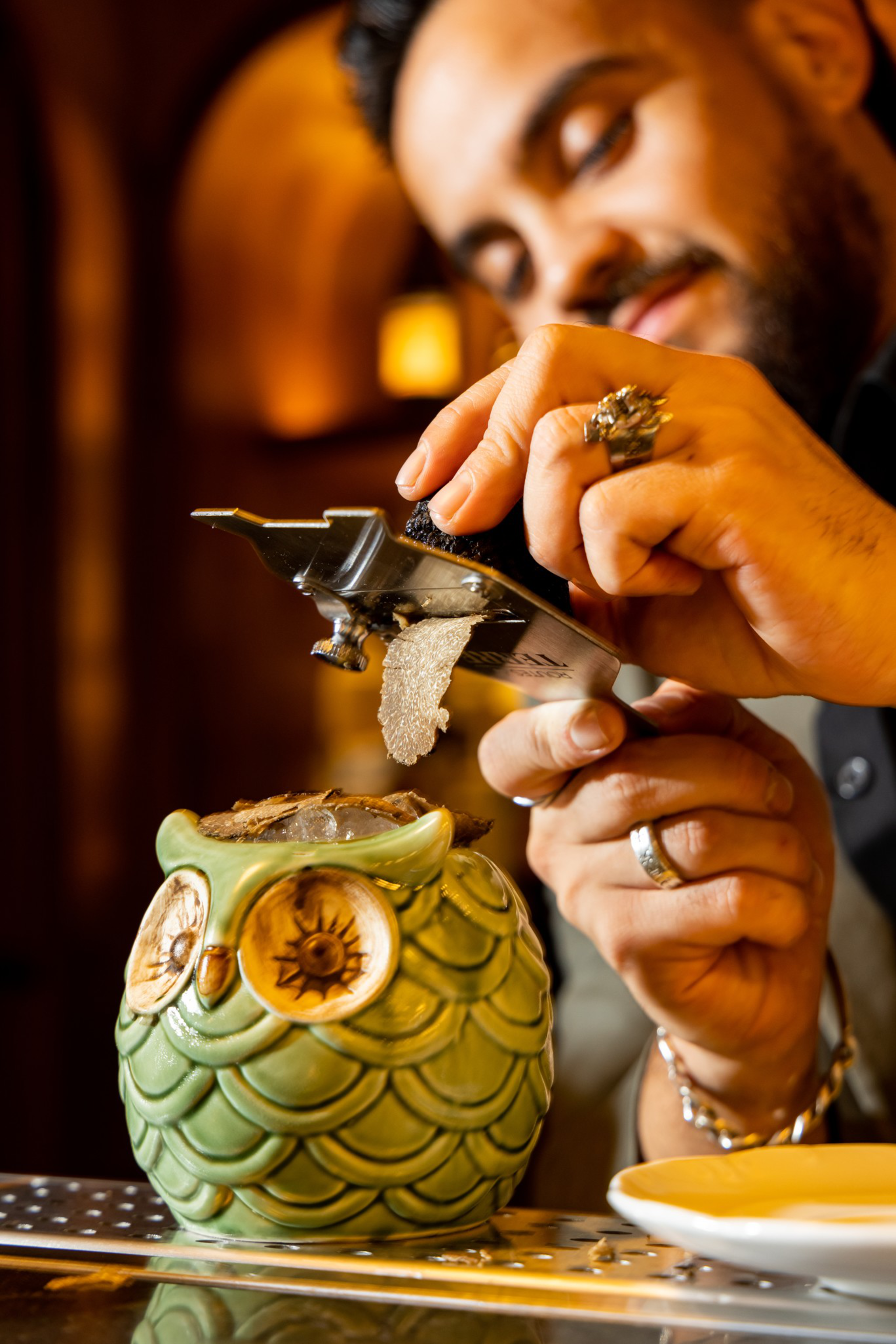 A man grates a piece of truffle over a green ceramic owl-shaped cup filled with a drink, focusing intently on his task.