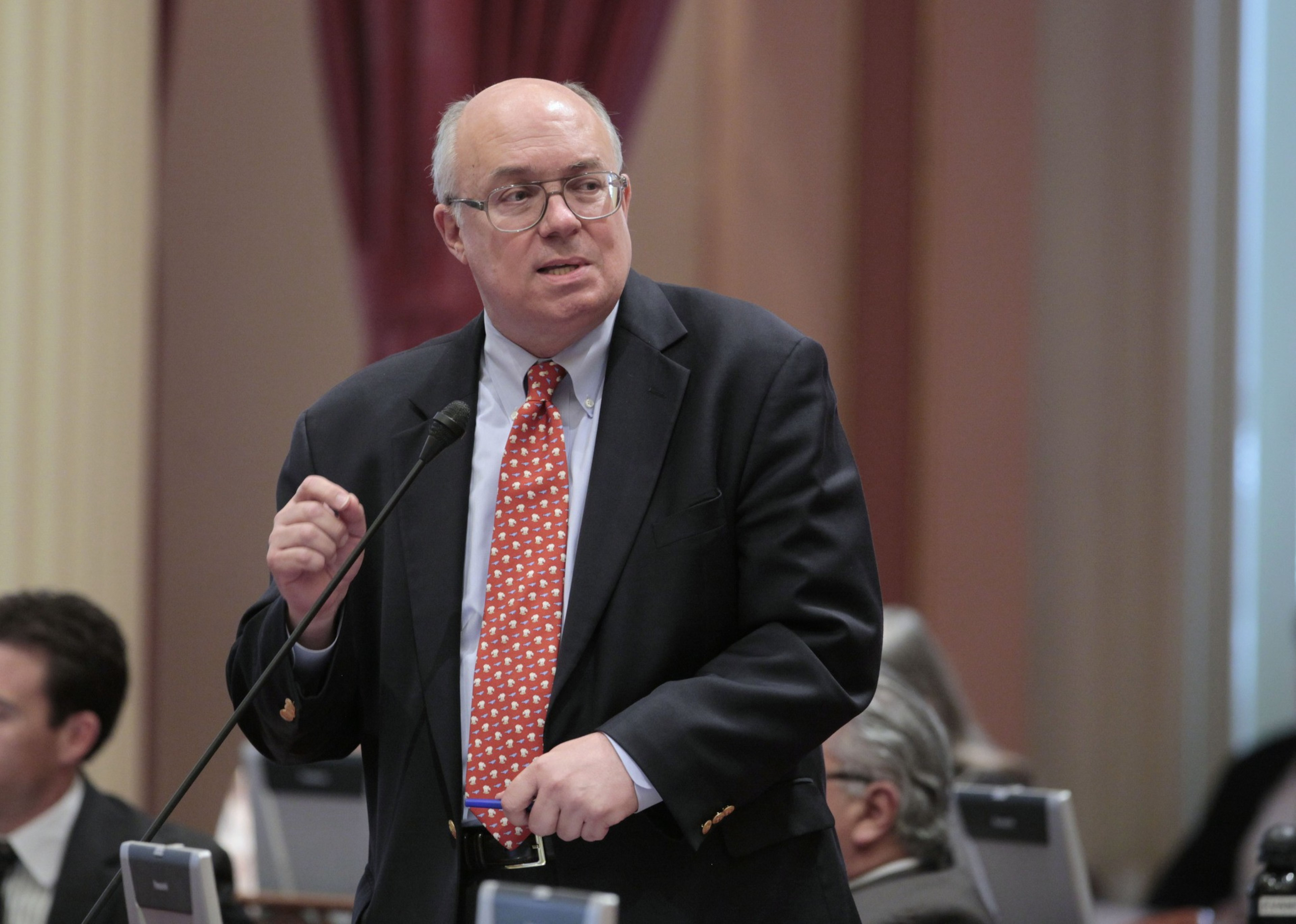 A man in a suit speaks at a podium with microphones, wearing glasses and a patterned tie.