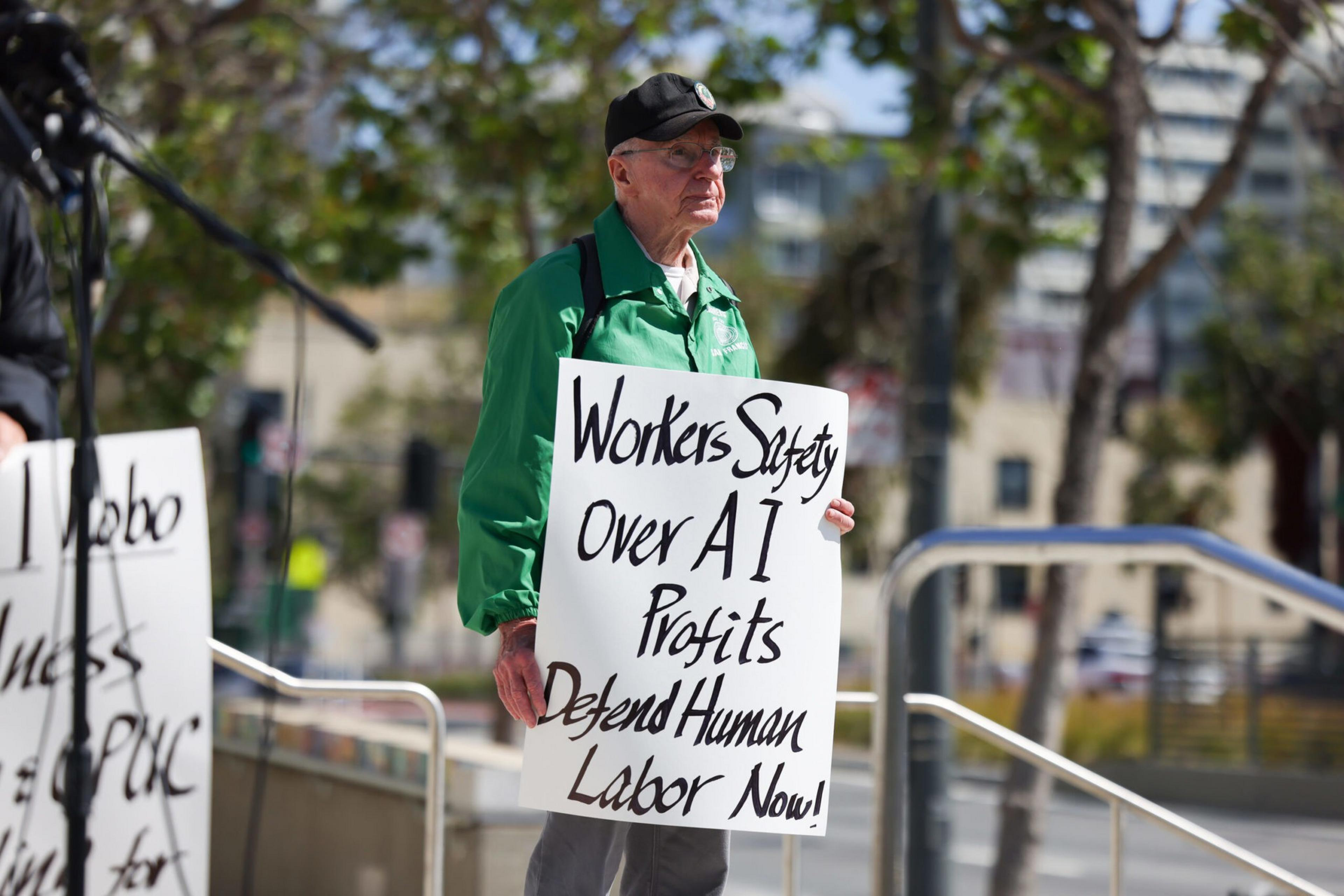 An elderly man in a green jacket and black cap holds a sign reading "Workers Safety Over AI Profits Defend Human Labor Now!" outdoors.
