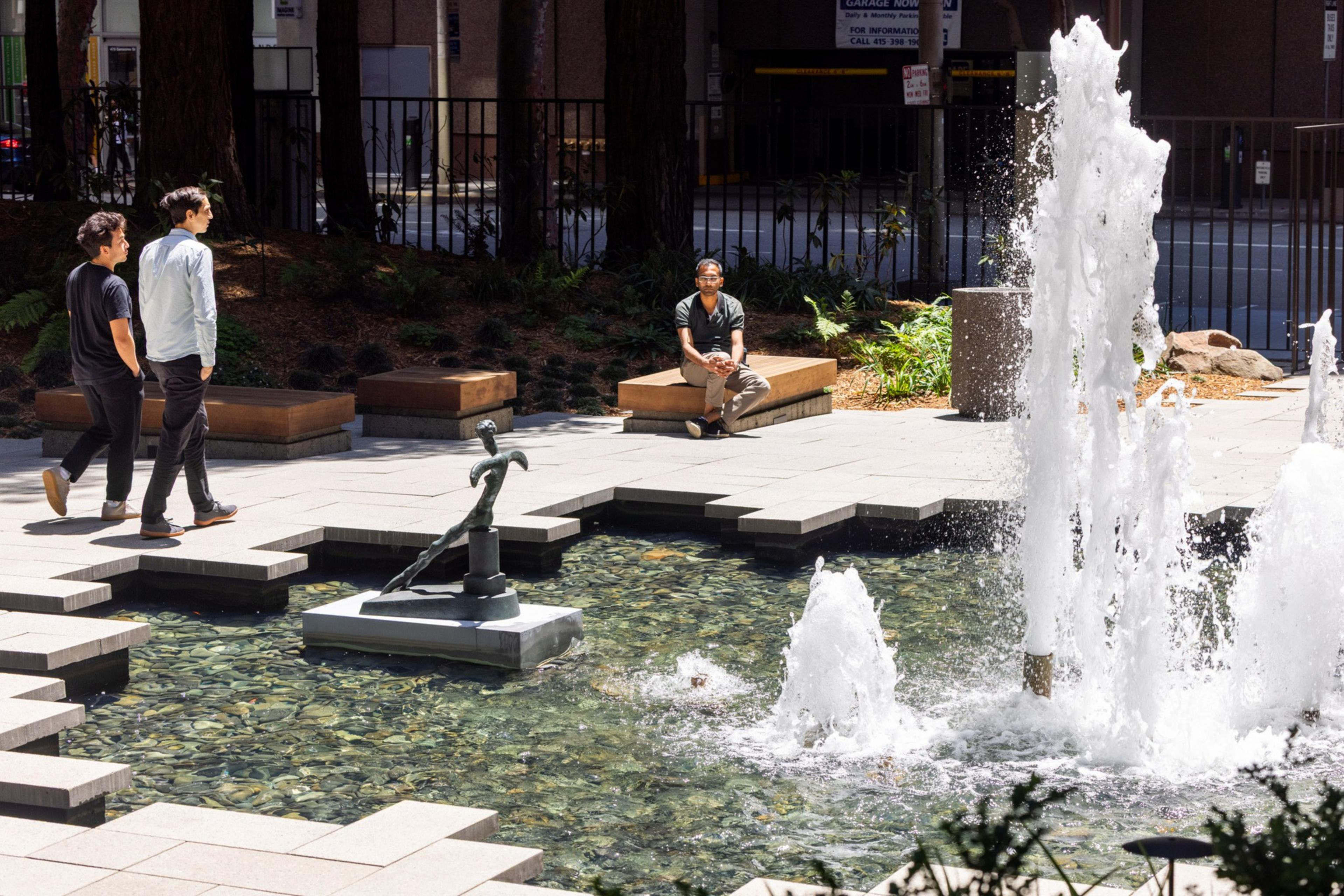 A small plaza features a fountain with a sculpture in a pool. Two people walk by, while another person sits on a bench in the sunlit, tree-lined area.