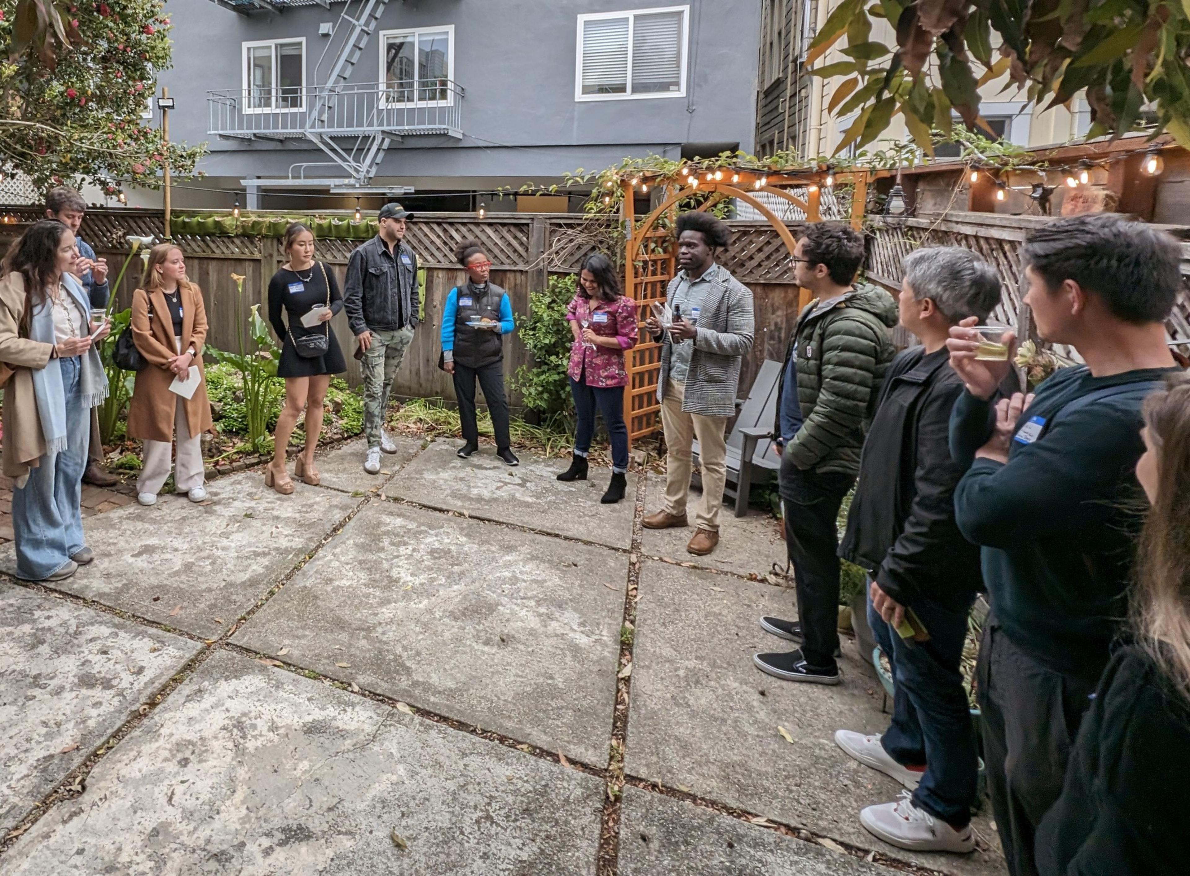 A group of around fourteen people stands in a circle in a backyard with a wooden fence and a trellis adorned with string lights. The setting is casual and conversational.