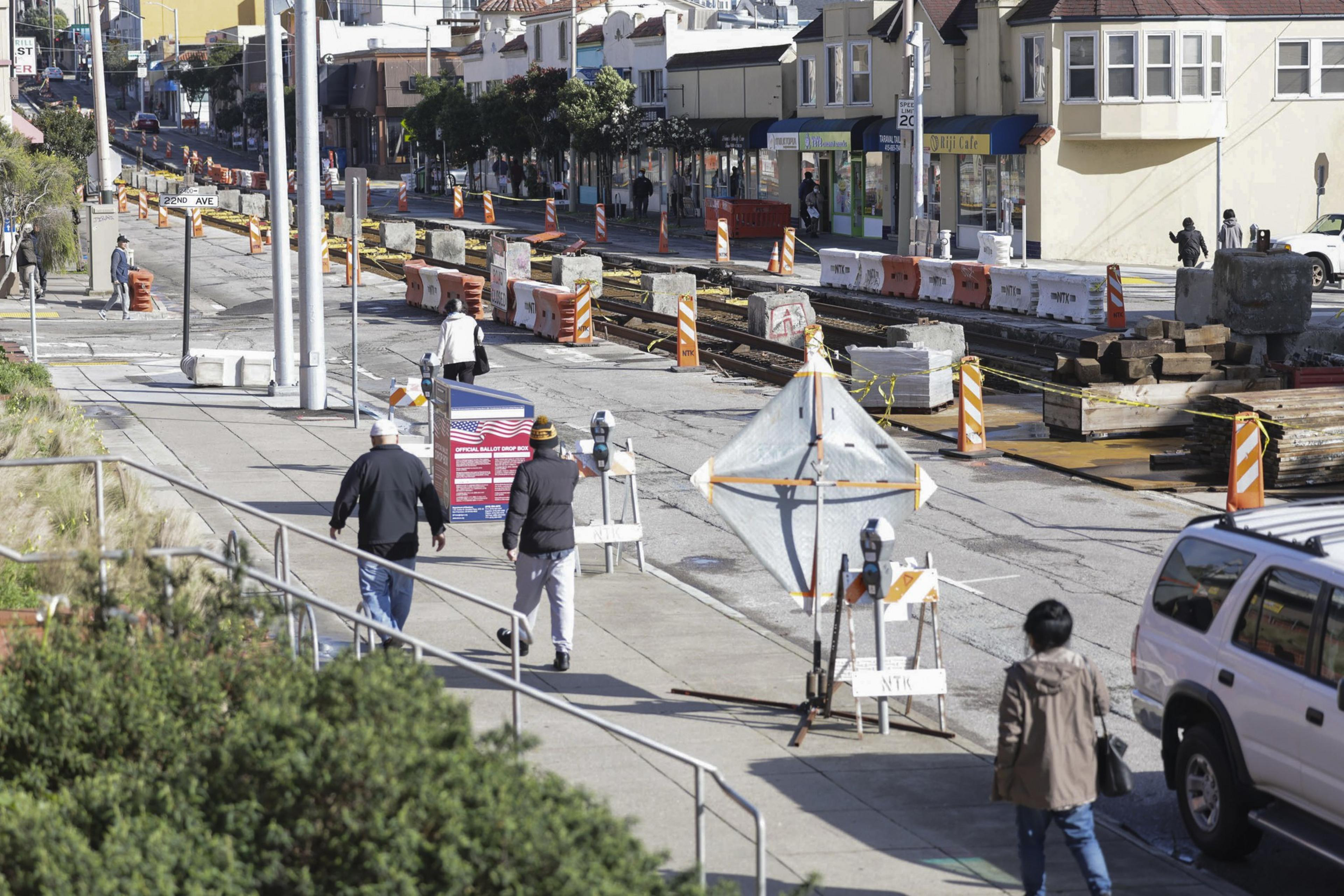 A busy street under construction with pedestrians, equipment, and an official ballot drop box.