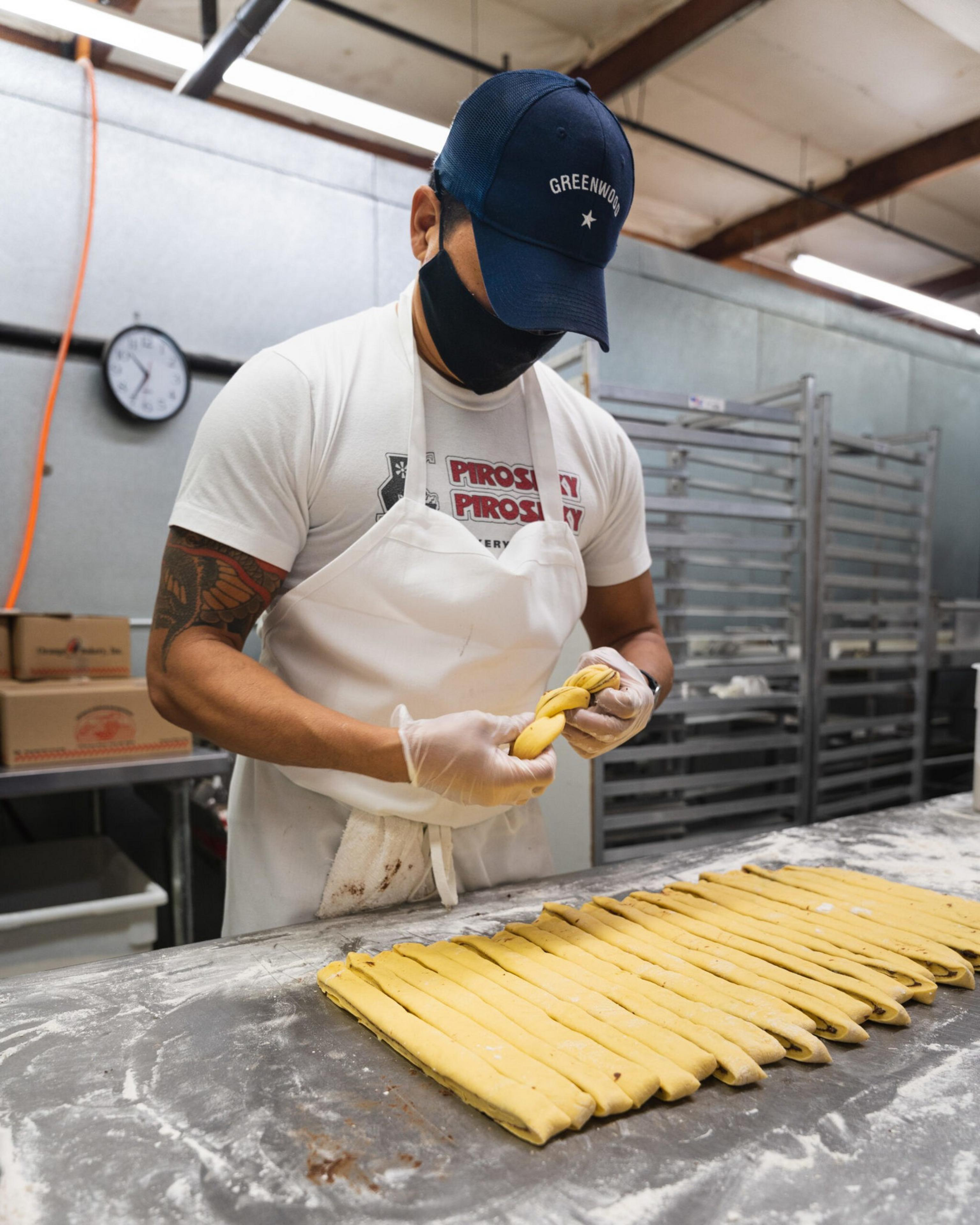 A person wearing a "Greenwood" cap, white apron, and gloves braids strips of dough on a flour-dusted table in a bakery setting.