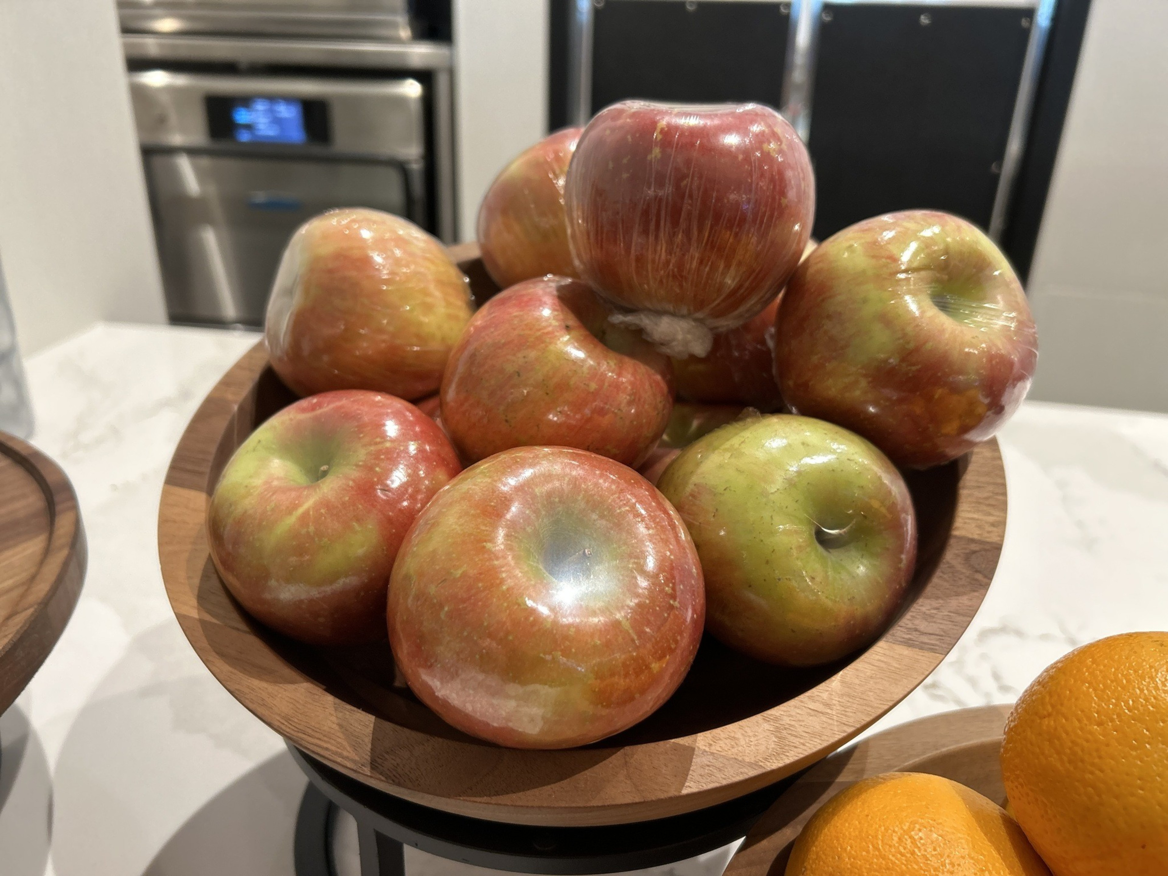 A wooden bowl is filled with shiny, red and green apples, some wrapped in plastic, placed on a kitchen counter near an oven.