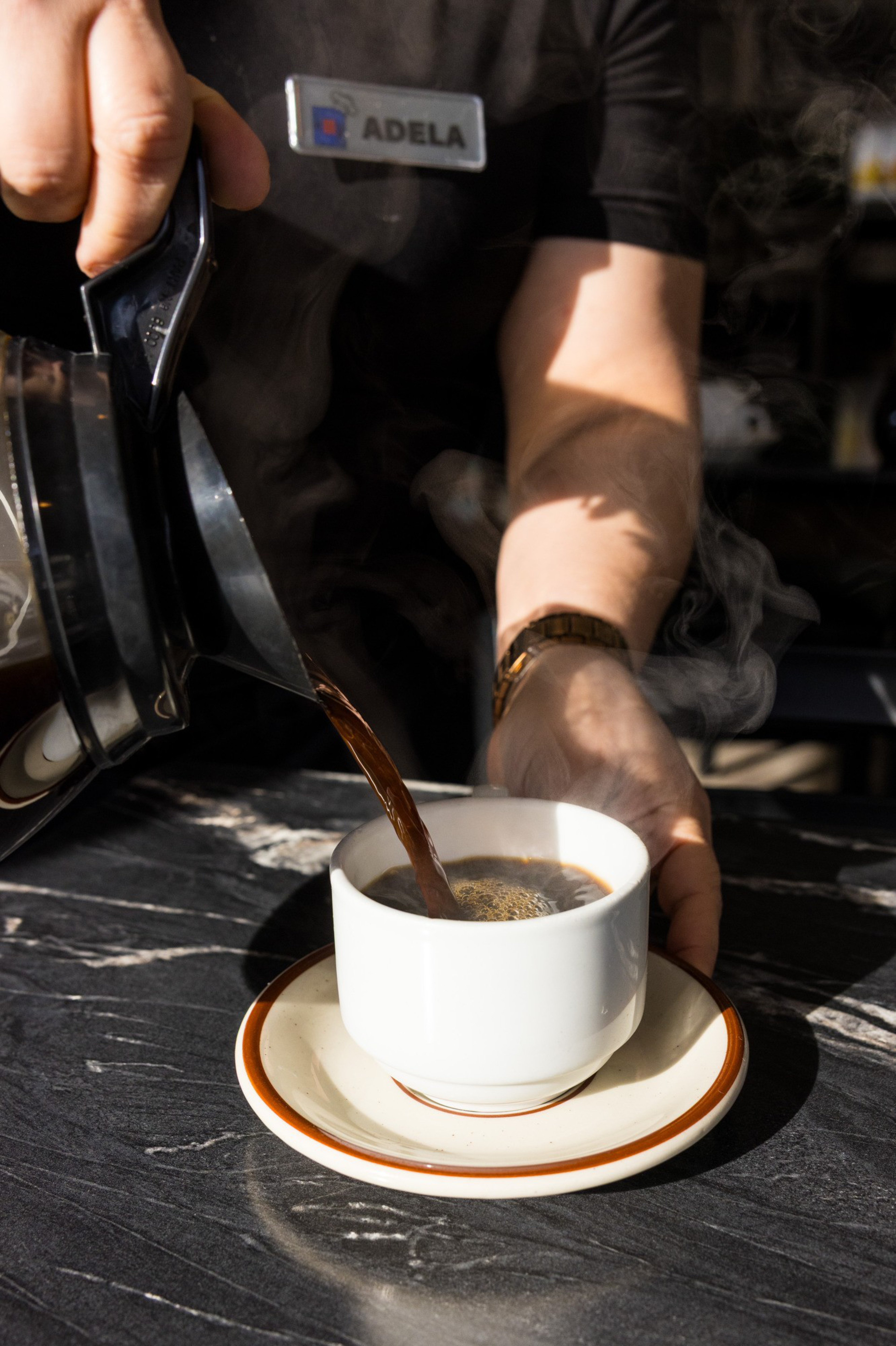 A person pours steaming coffee from a pot into a white cup on a saucer, placed on a dark marble surface. They're wearing a name tag that says &quot;Adela.&quot;