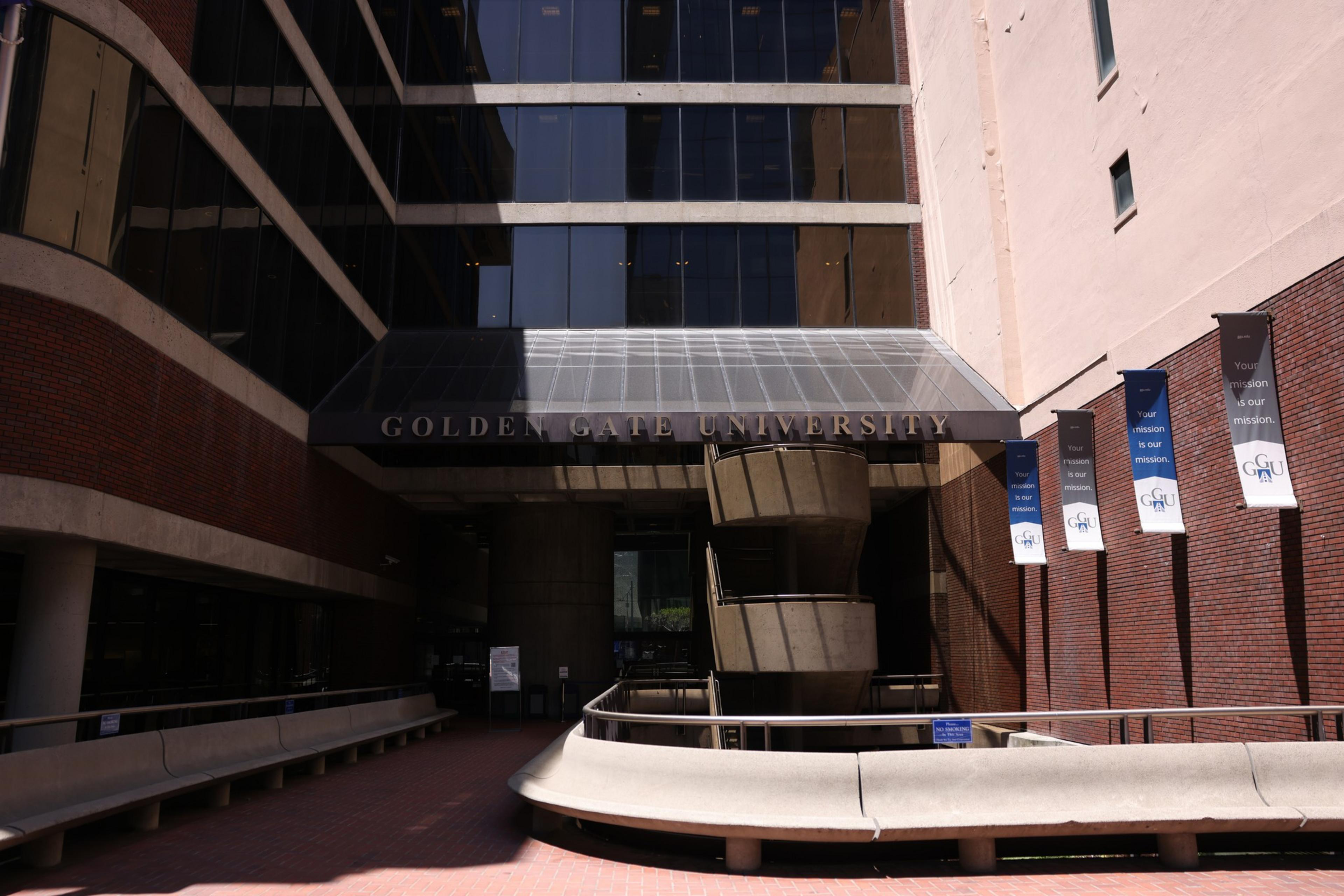 A modern university entrance with reflective glass windows, a canopy reading "Golden Gate University," curved concrete benches, and hanging banners on a brick wall.