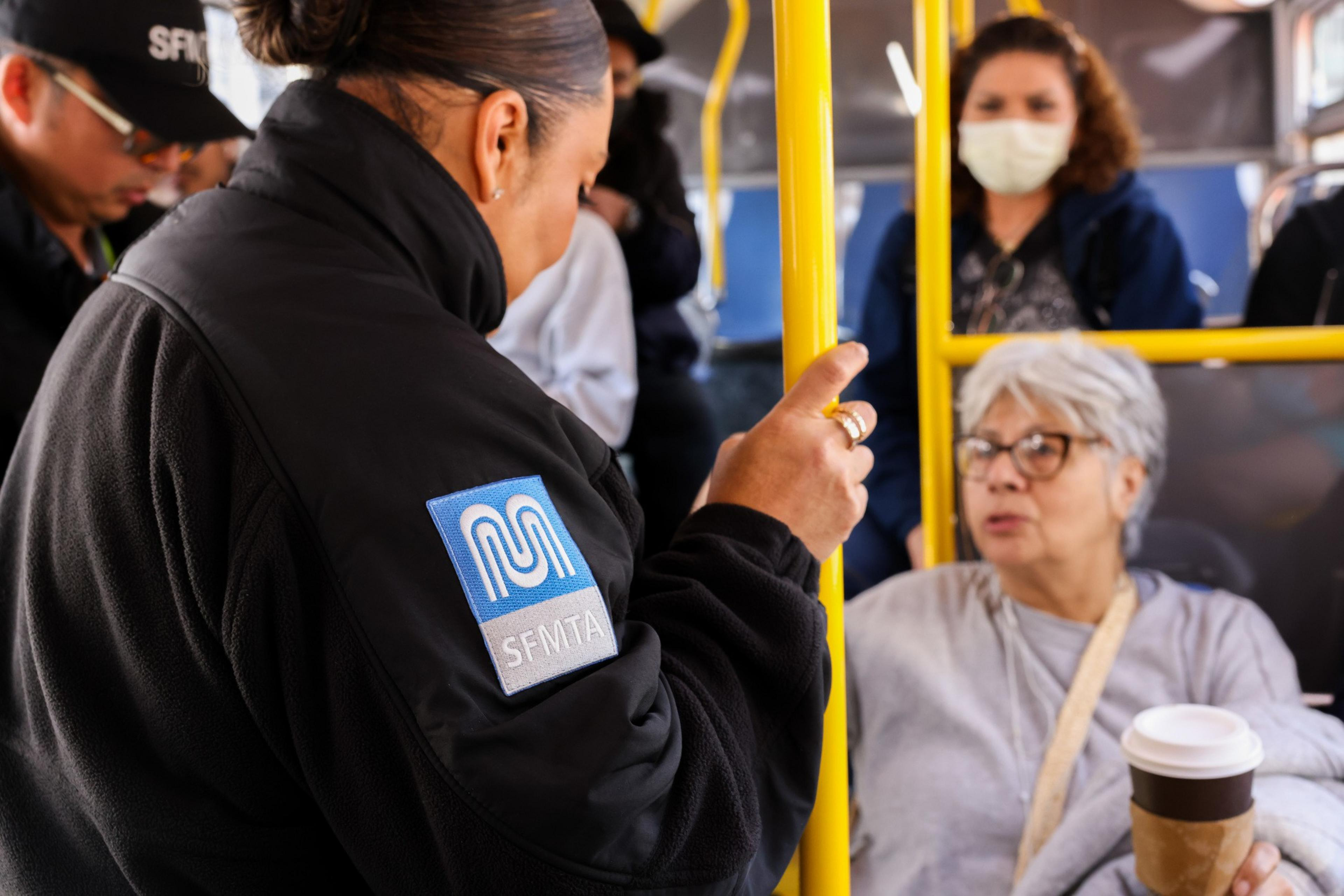 A transit officer speaks with an older woman holding a coffee on a bus. Other passengers, including a mask-wearing person, are in the background.