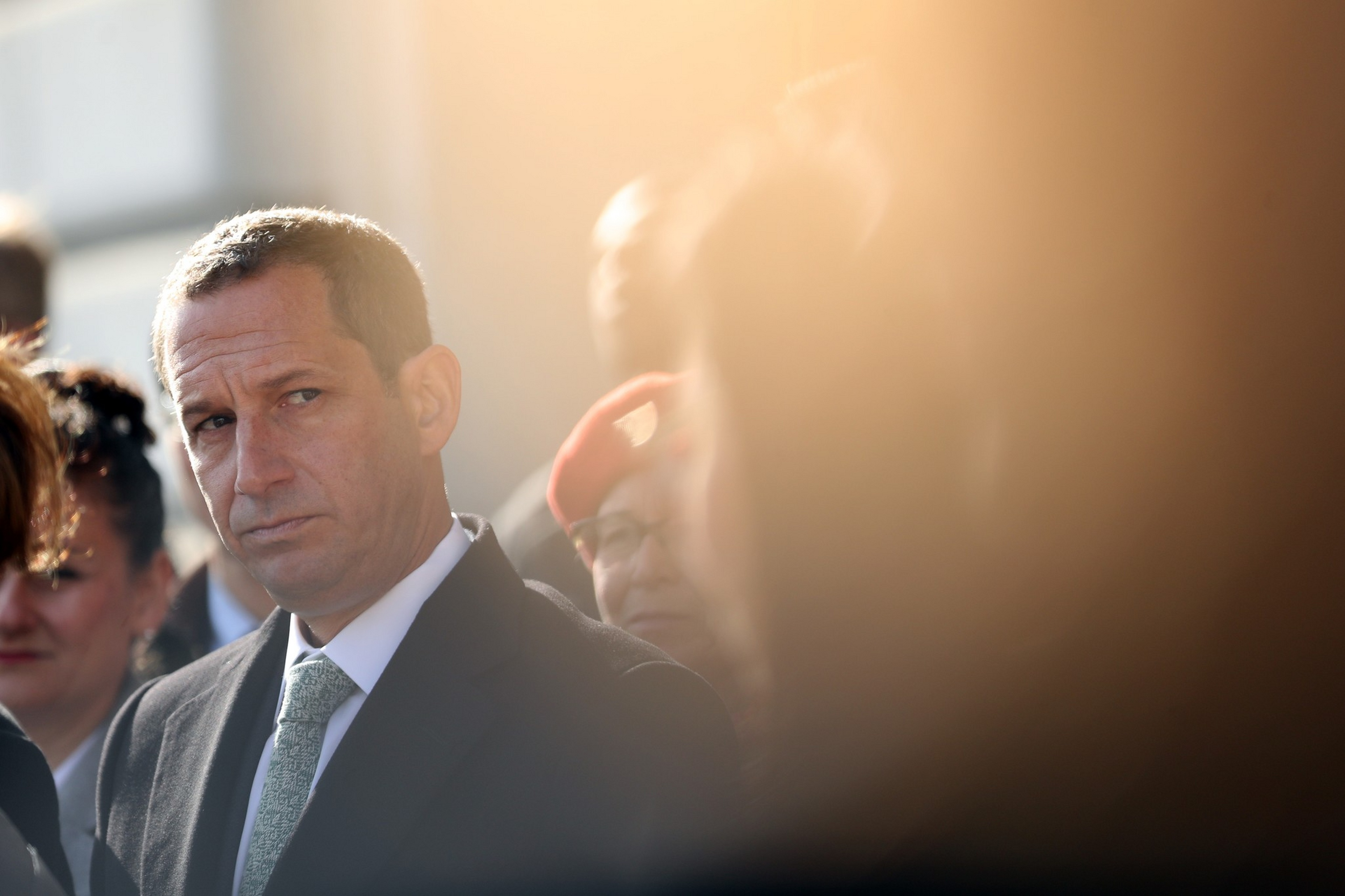 A man in a suit and tie looks thoughtfully to the side, surrounded by a blurred crowd with soft sunlight creating a warm glow.