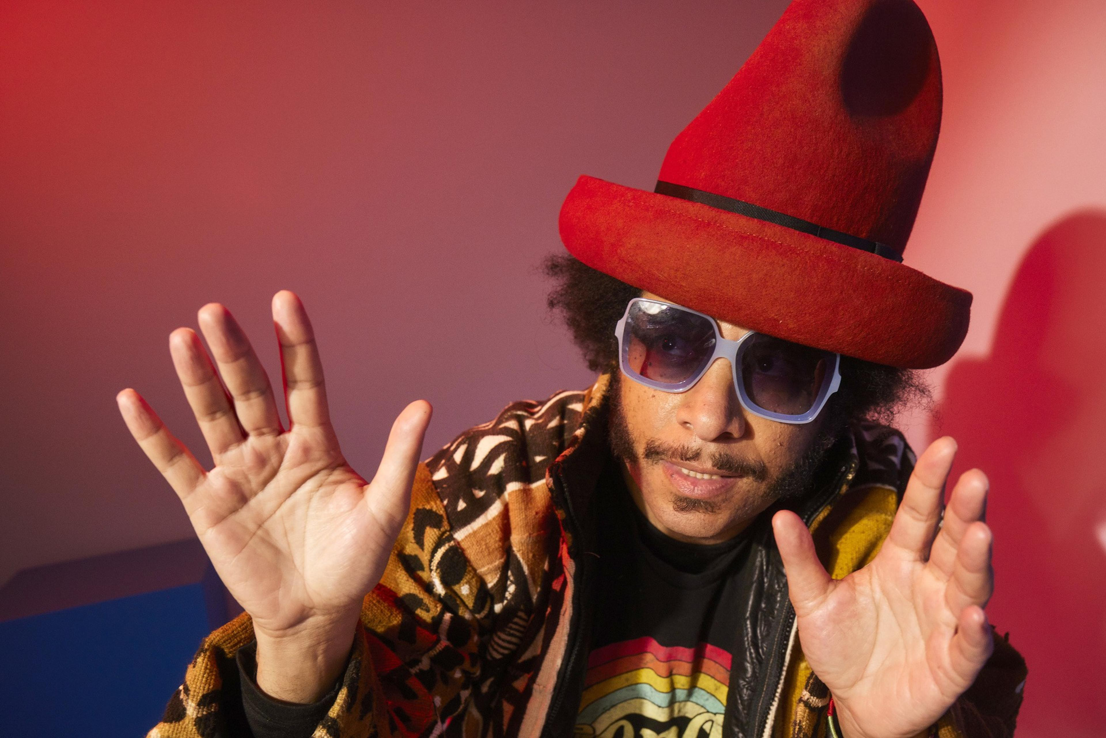 A person wearing a large red hat, white-framed sunglasses, and a patterned jacket raises both hands toward the camera against a red background.