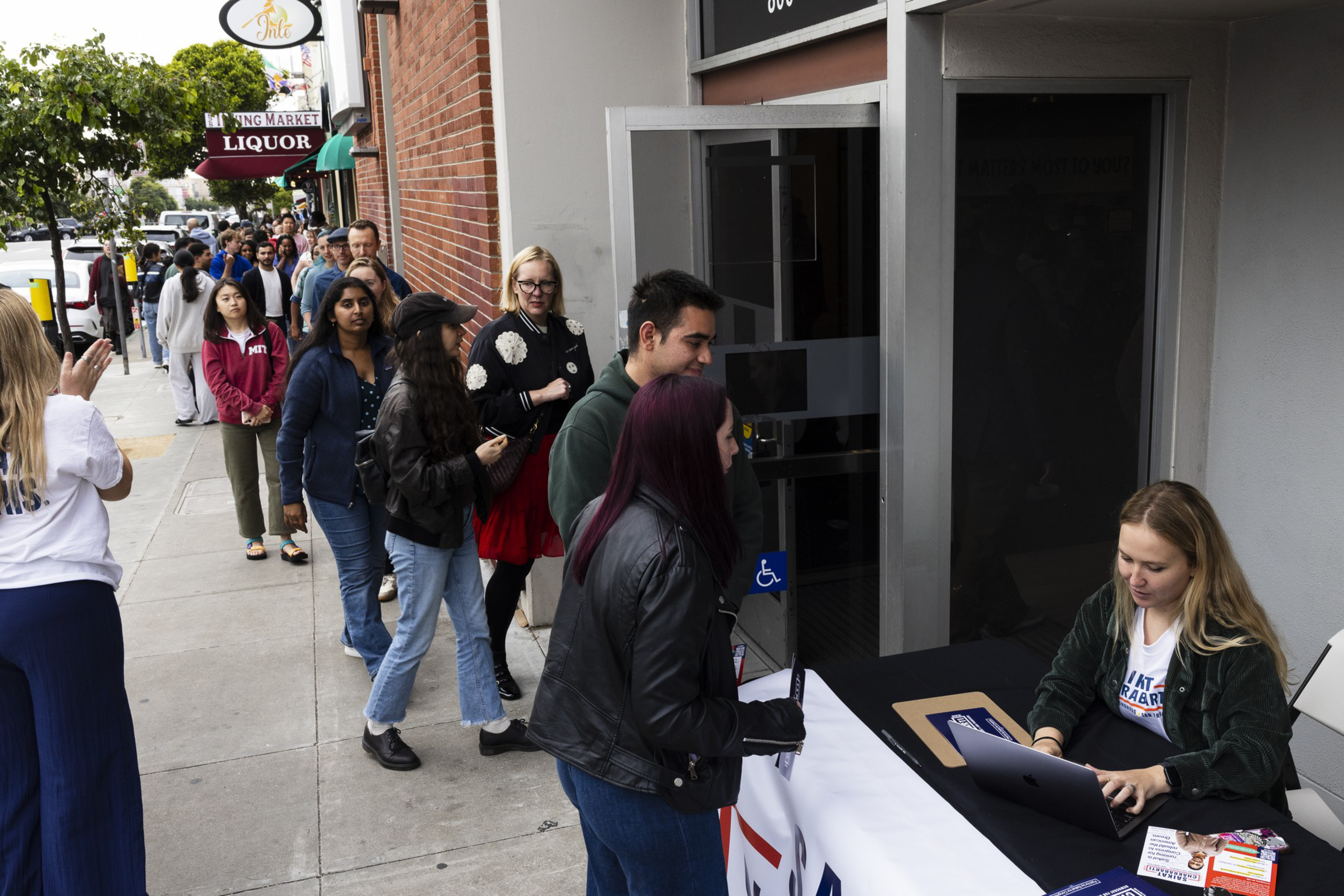 People wait in a long line along a sidewalk that leads to a doorway where a woman seated at a table with a laptop assists those arriving.