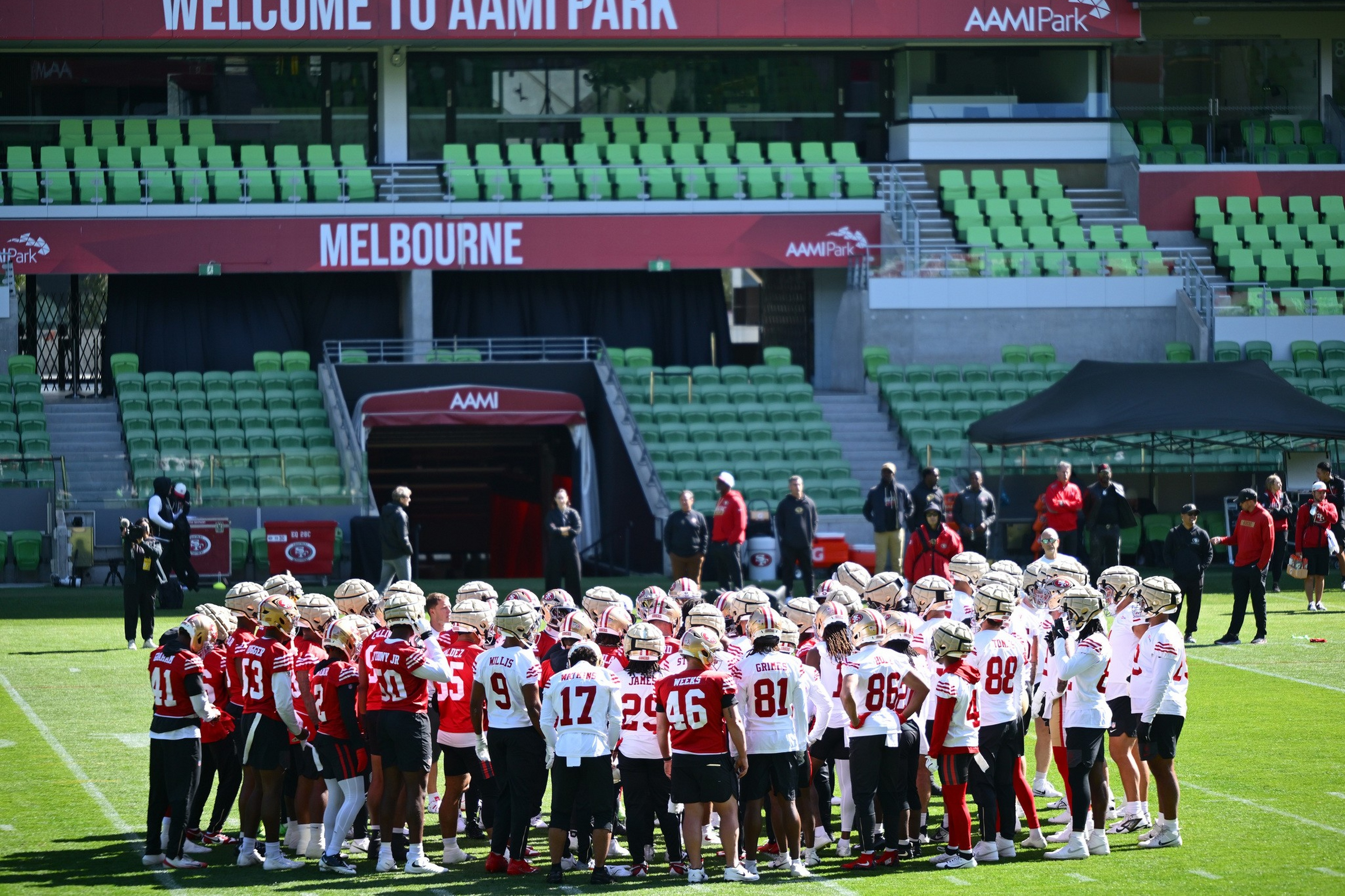 A large group of football players in red and white uniforms huddle together on a green field at AAMI Park, Melbourne, with empty green stadium seats in the background.
