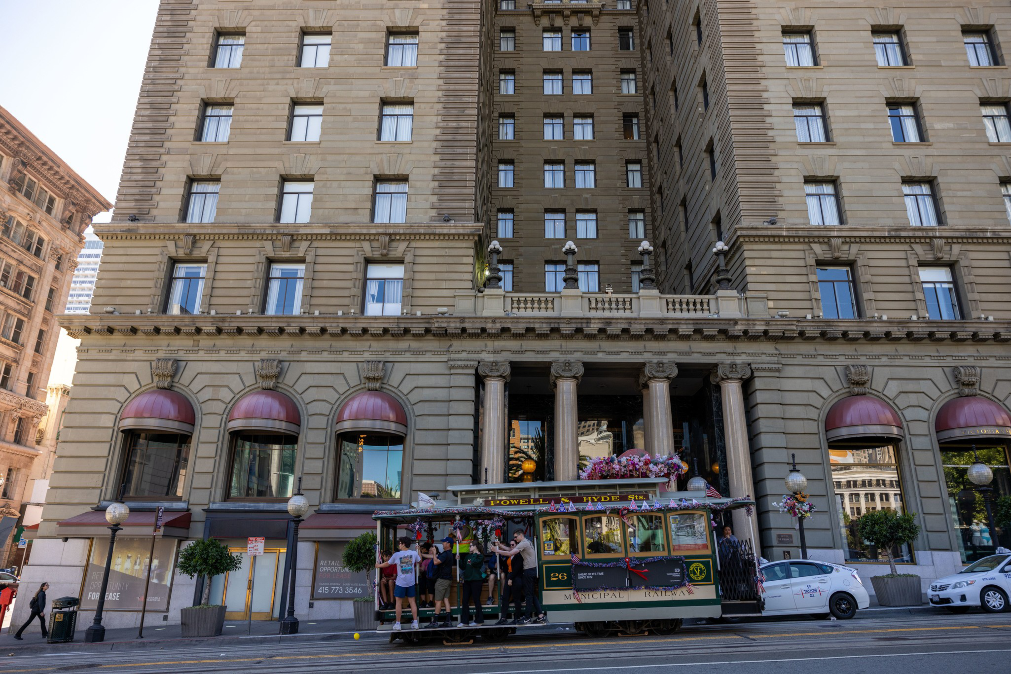 A decorated cable car filled with people passes in front of a historic building with columns. The scene includes a few pedestrians and parked cars.