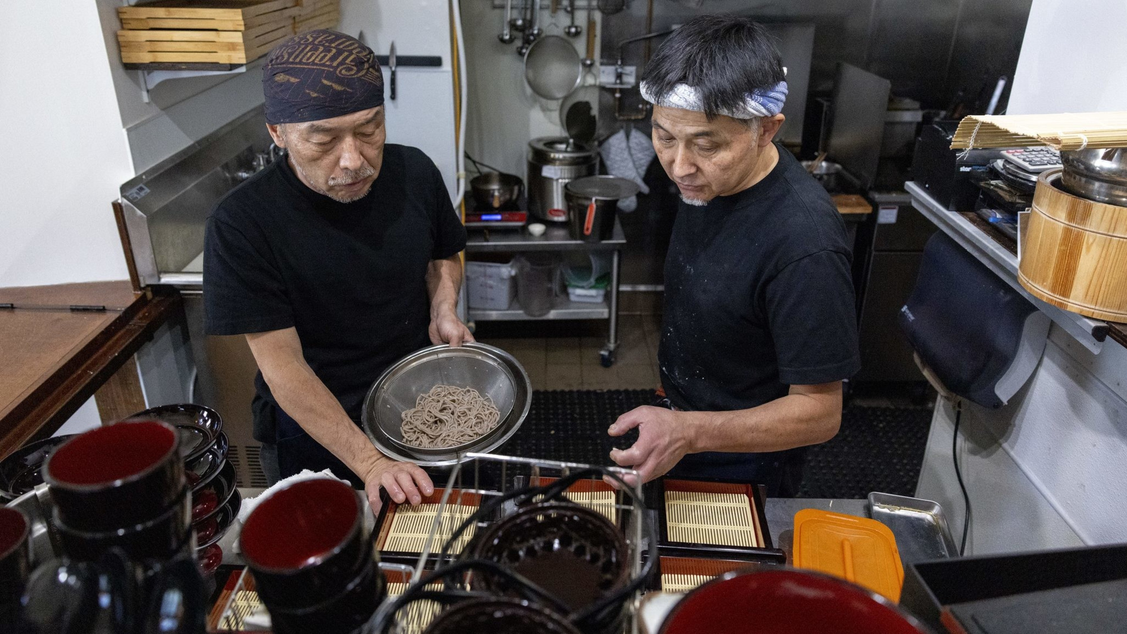 Two men in a kitchen prepare soba noodles. One holds a bowl of noodles while the other arranges dishes. The kitchen is filled with cooking utensils and dishes.