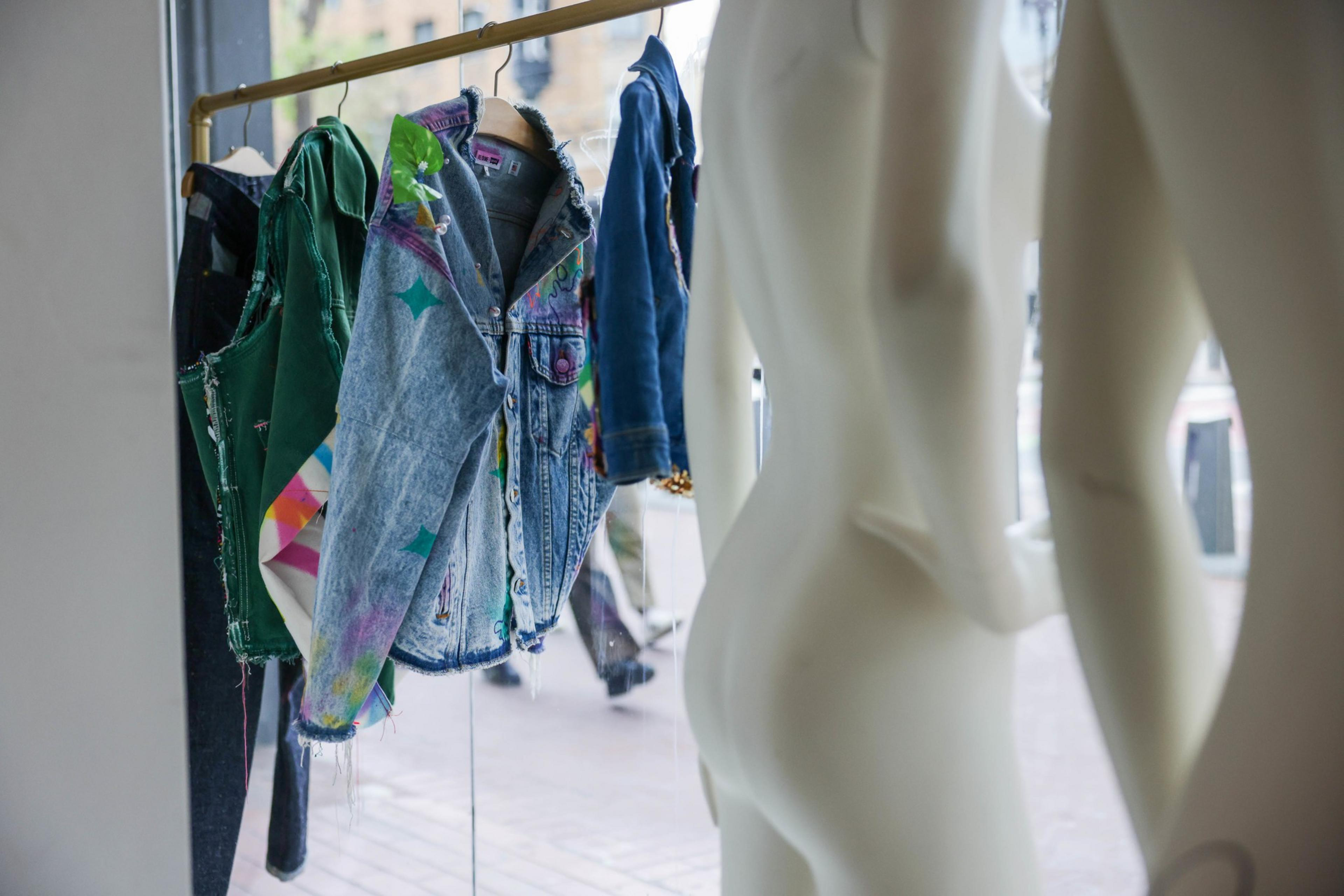 Colorful, distressed denim jackets hang on a rack, with a clear view through a window behind.