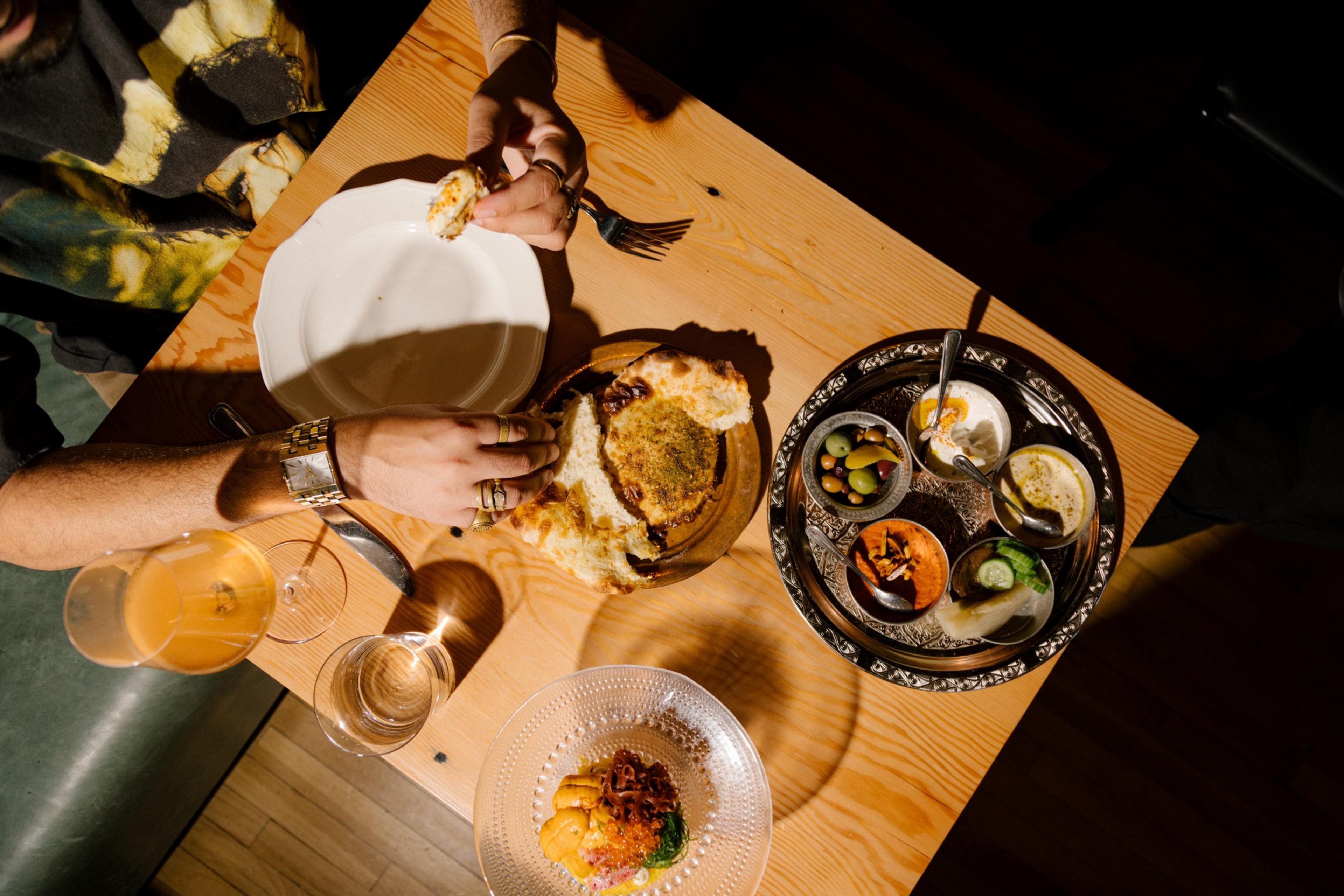 A overhead photo of a table with plates and serving dishes that include hummus, muhammara, smoked yogurt, pickles, marinated olives and pita with the hands of a person ripping pita bread before eating it.