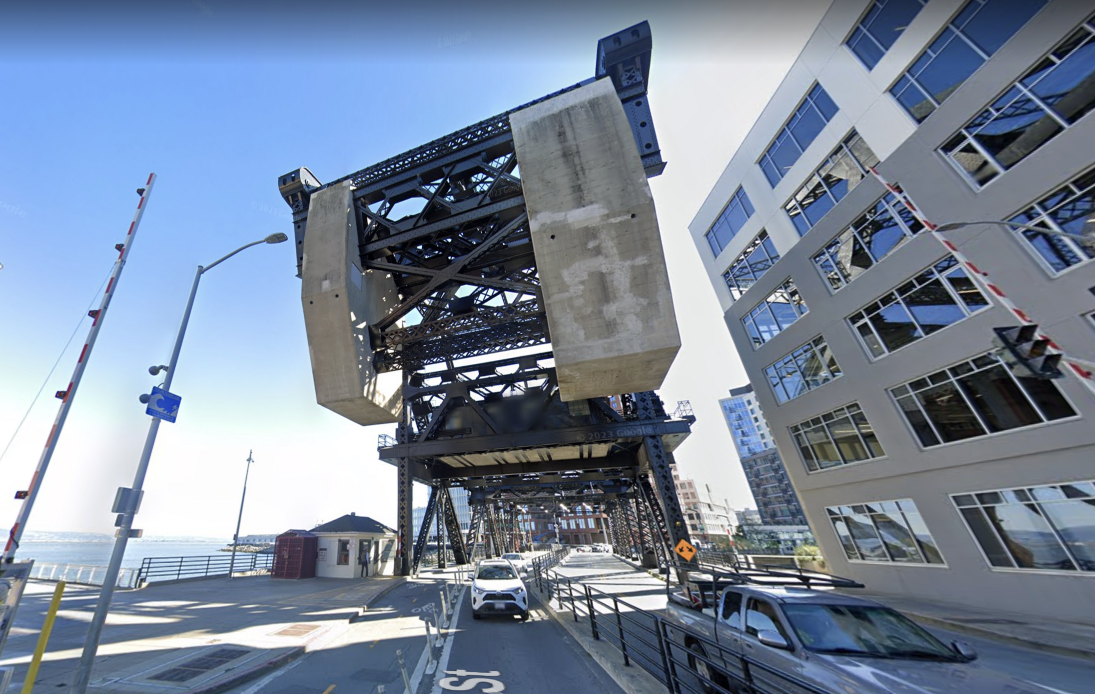 Cars pass under a large metal bridge with a commercial building to the right. 