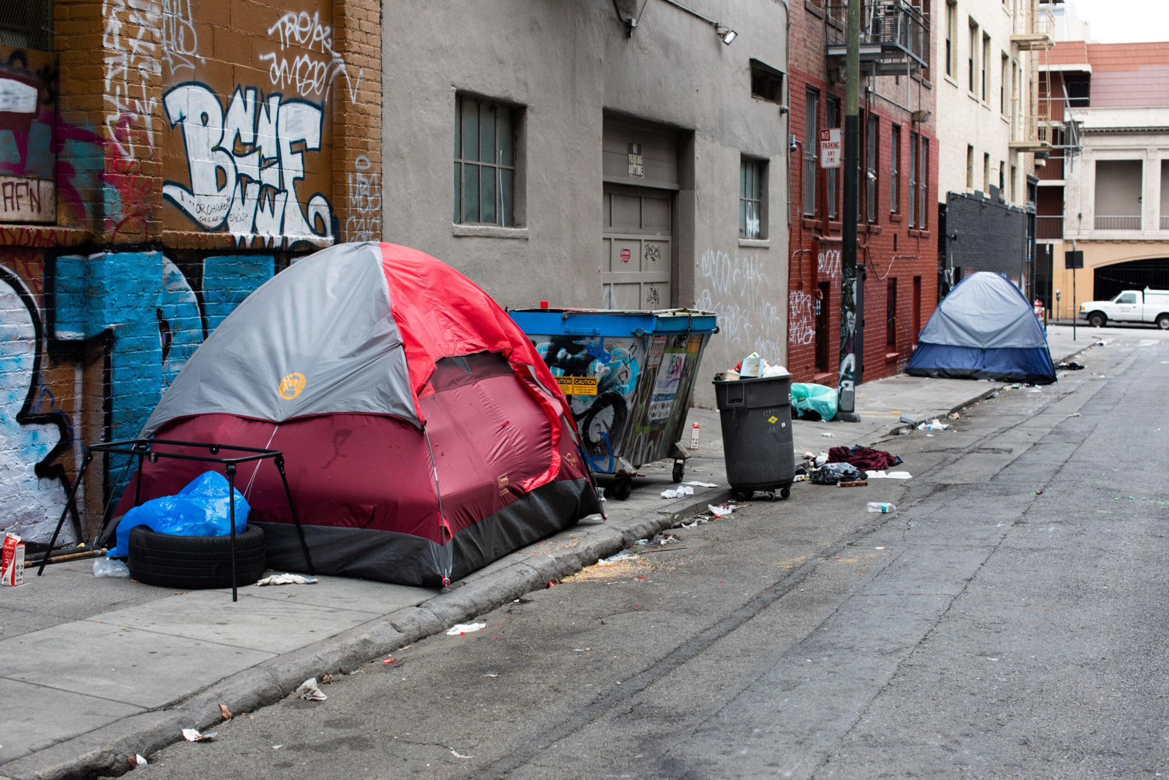 Two tents are set up on a graffiti-covered urban sidewalk next to a dumpster, with scattered trash and debris around them.