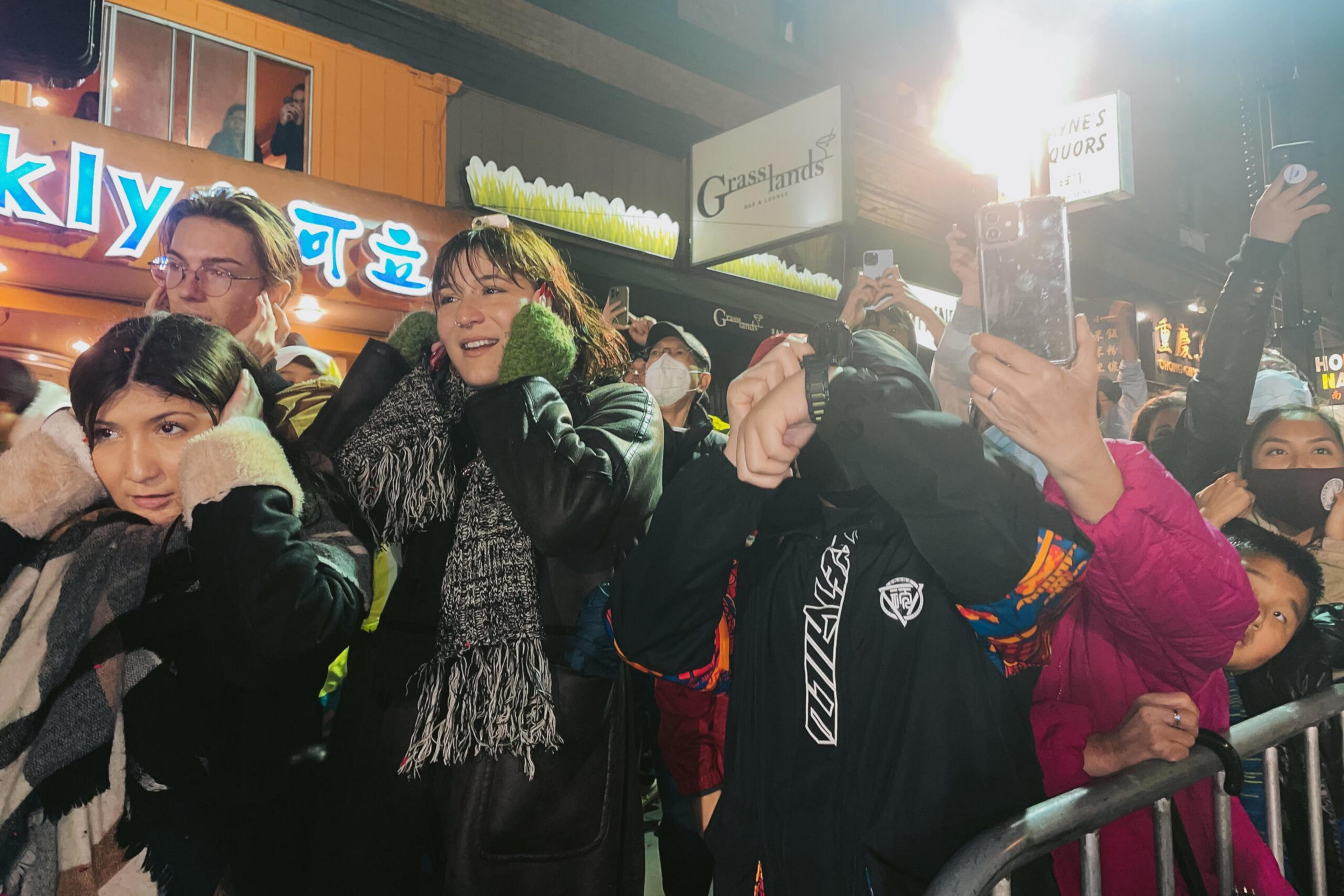A crowd of people bundled in winter clothes stand tightly together, some holding phones up to capture or record something off-frame.
