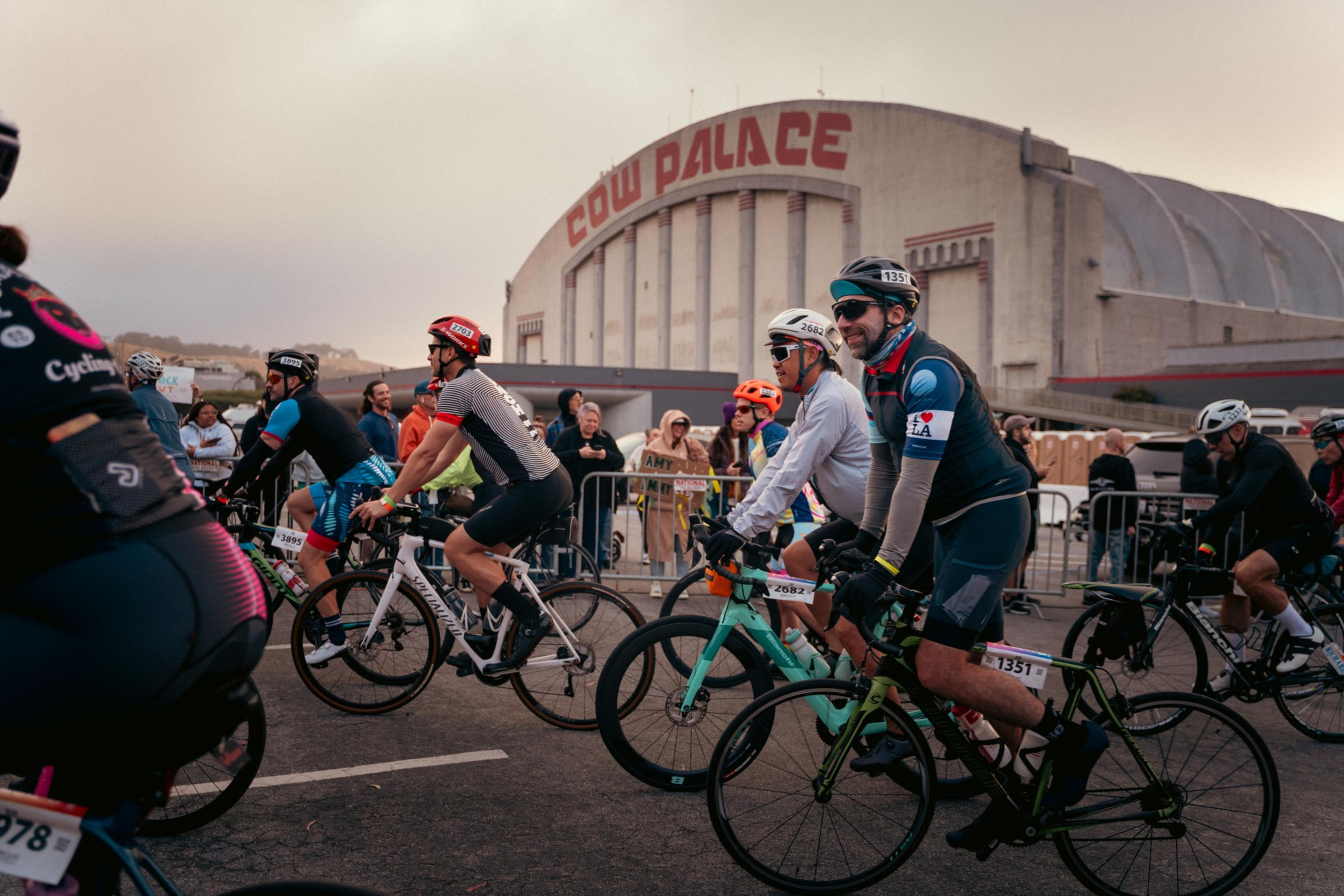 Cyclists in colorful gear ride past Cow Palace. Spectators cheer from behind barriers on a cloudy day, with one holding a sign for &quot;Amy.&quot;