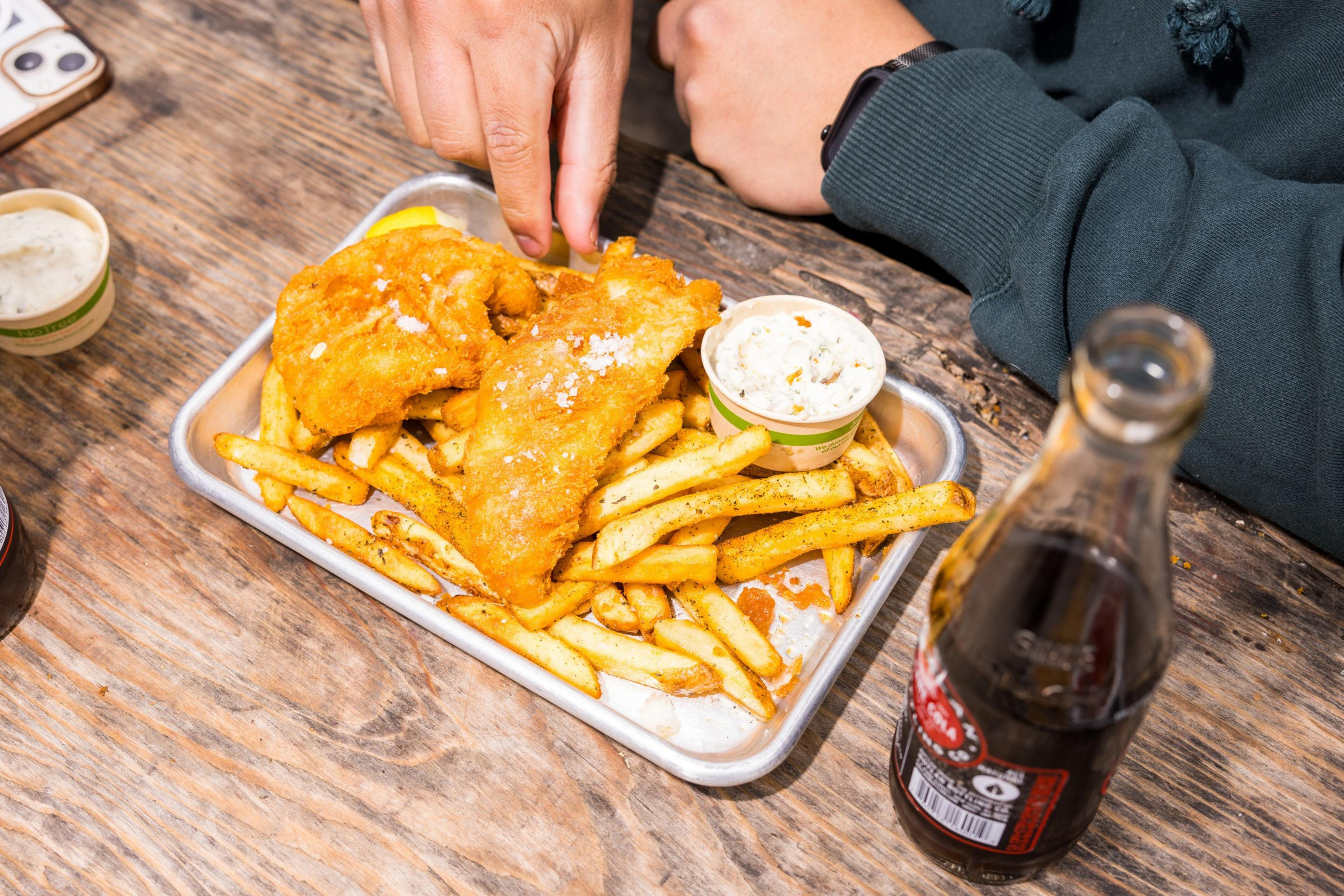 A person is enjoying a meal of fried fish and chips placed on a metal tray. There's a small cup of sauce and a bottle of cola on a wooden table.