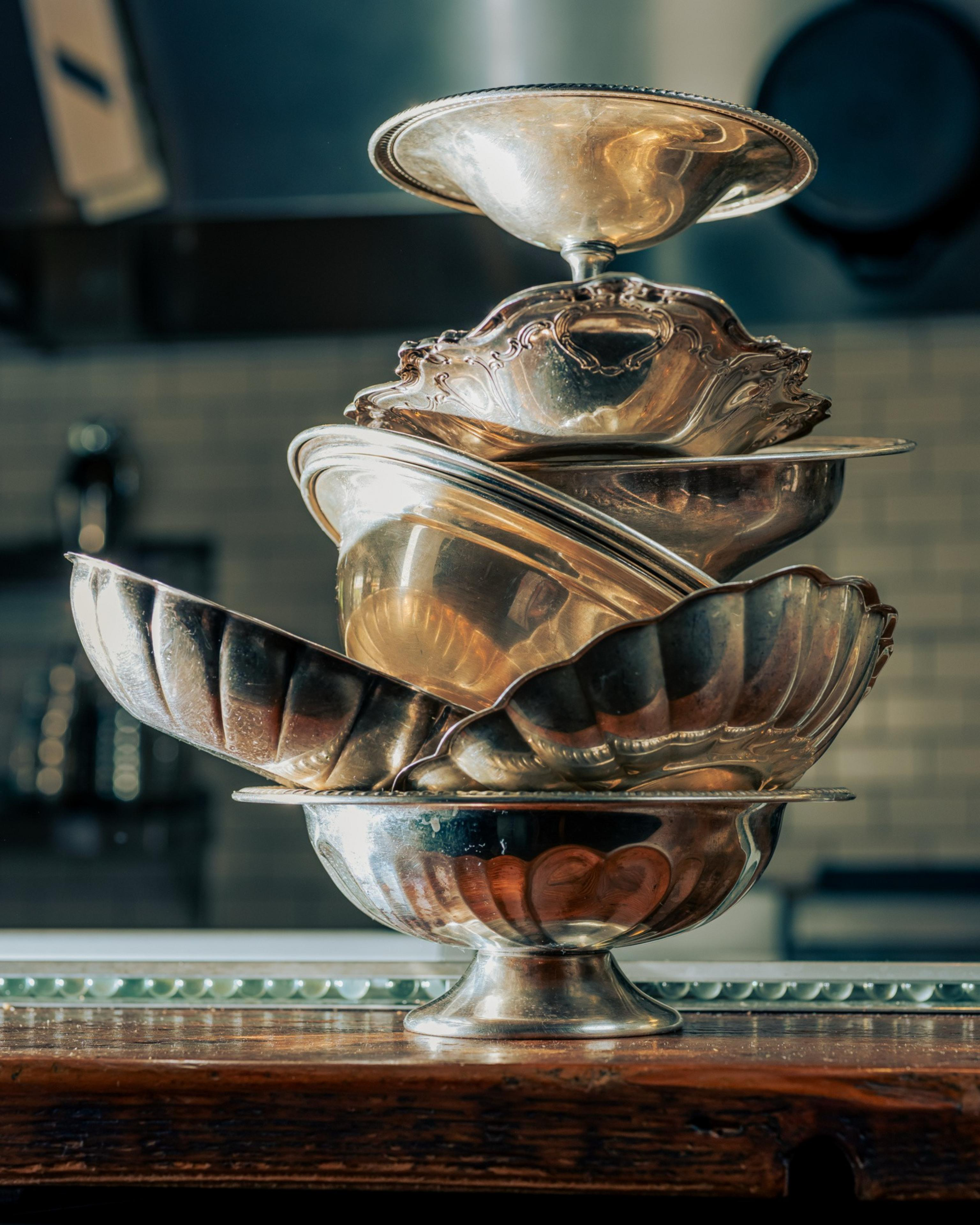 A group of shiny, stacked silver bowls of various shapes and designs sits atop a wooden surface, with a blurred kitchen background.