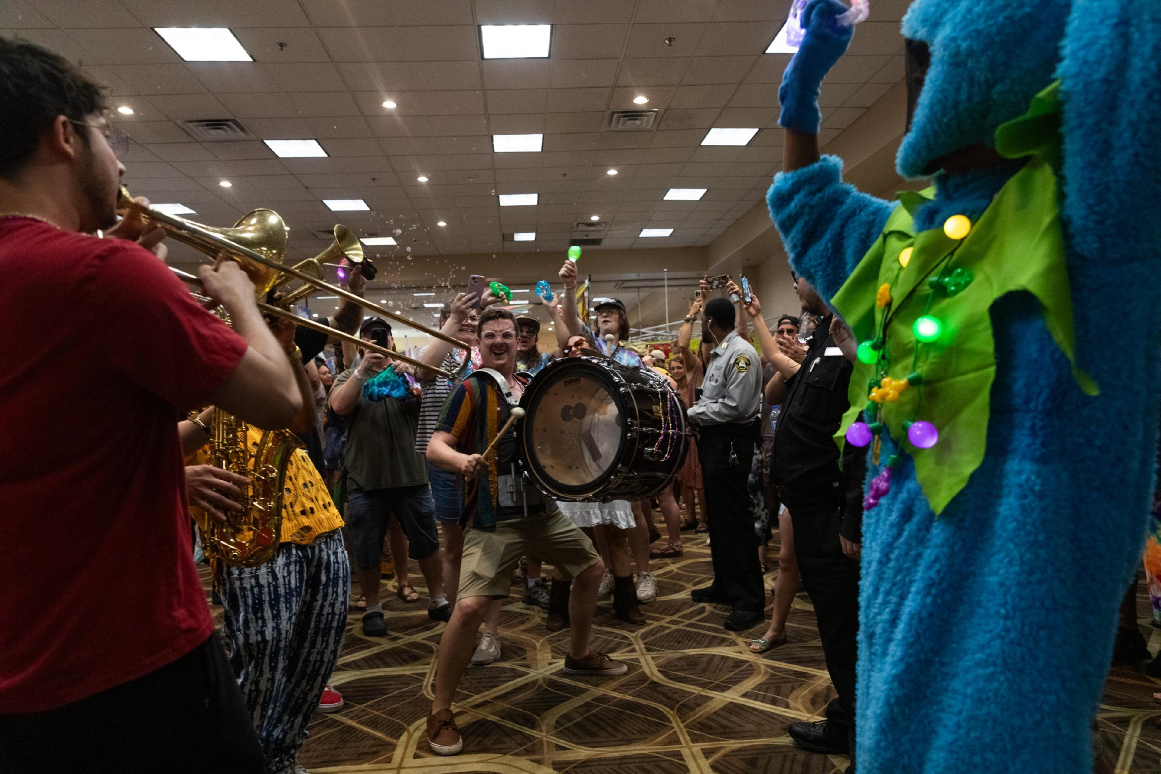 A lively indoor scene shows a costumed character in blue, illuminated by lights, dancing beside a band with brass instruments, and a cheering, colorful crowd.