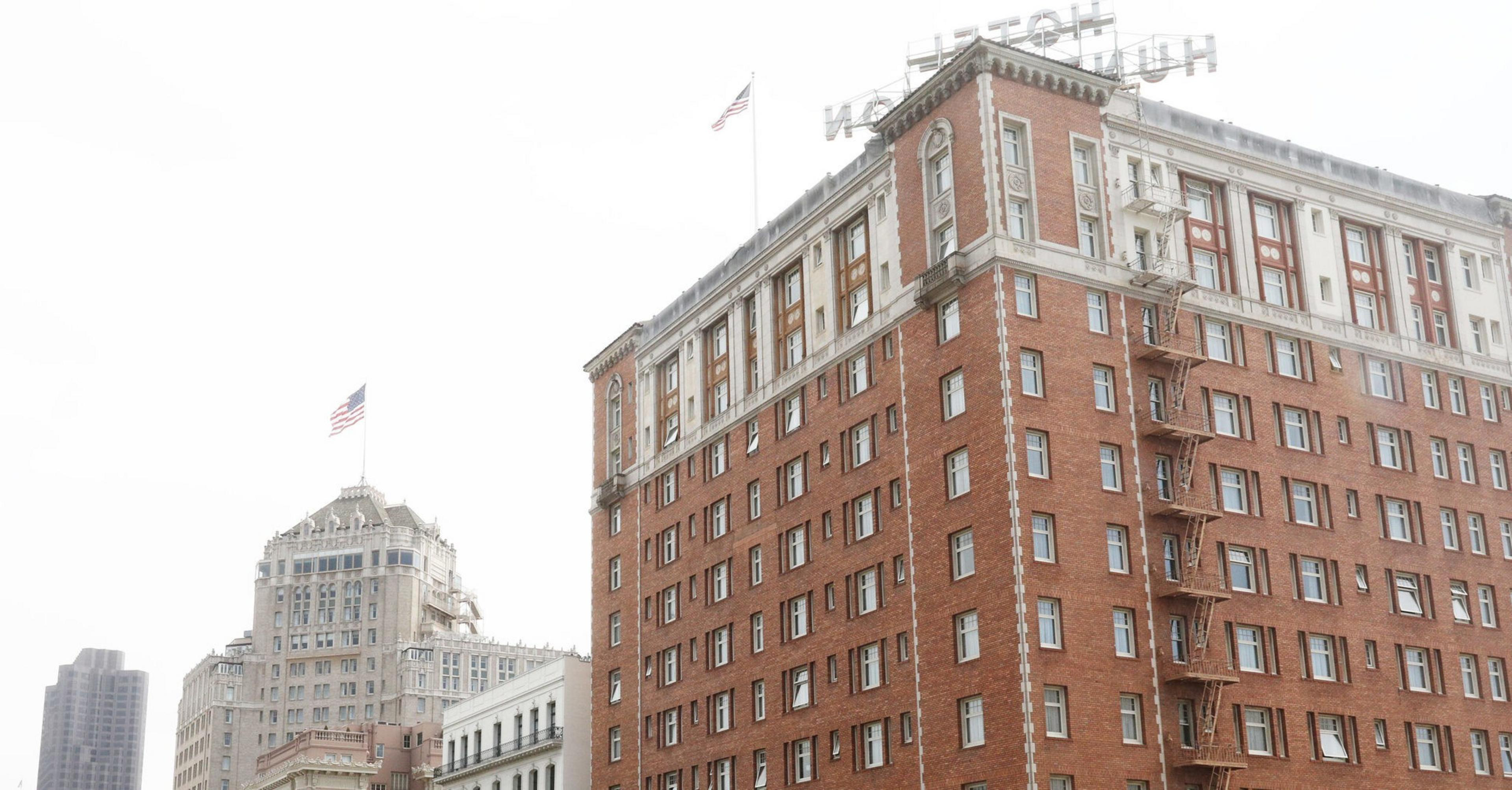 A red brick hotel building with many windows and fire escapes, topped with a flagpole flying an American flag, stands against a pale sky.