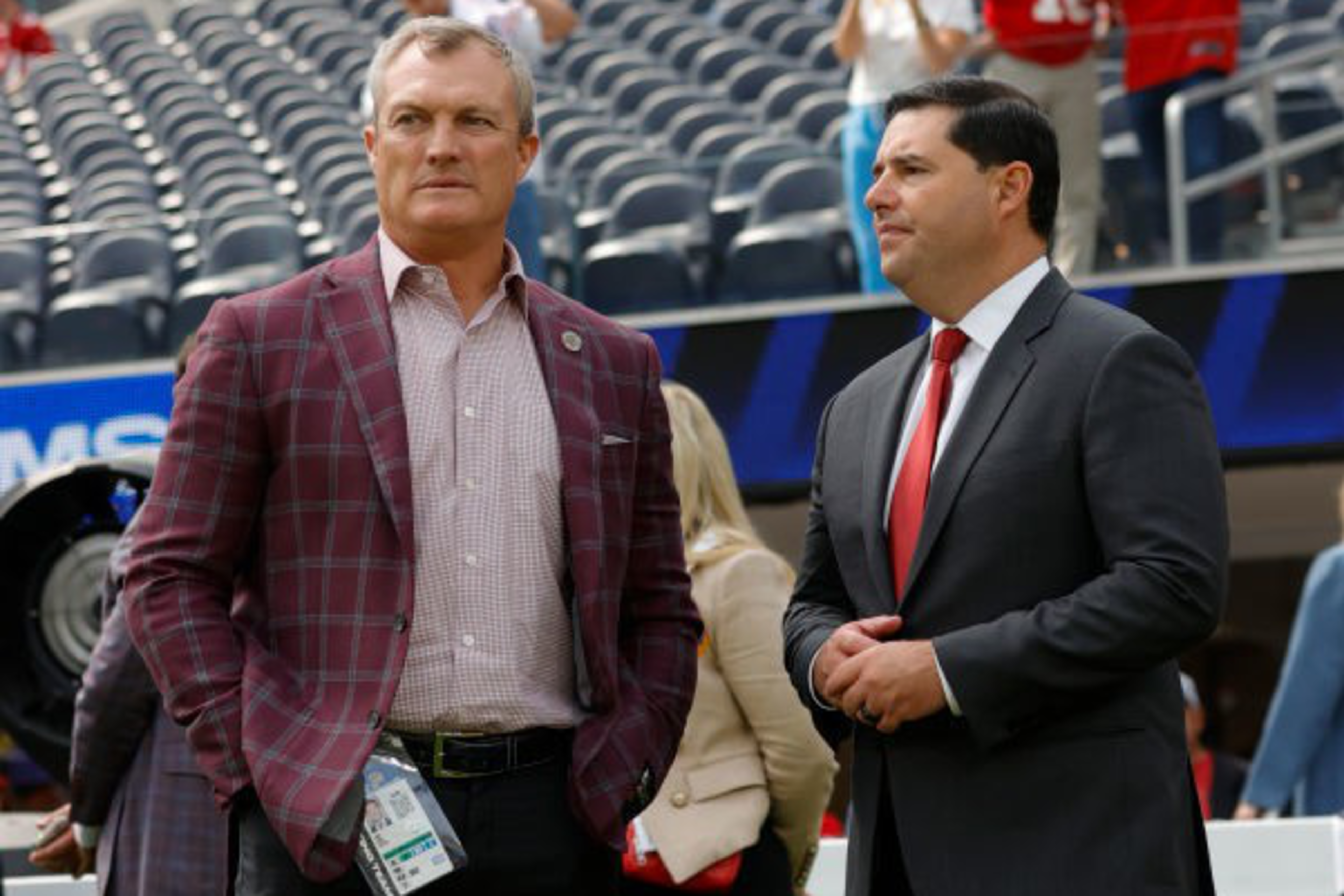 Two men in suits stand on a football field, with one wearing a plaid jacket and the other a red tie. People in the background occupy stadium seats.
