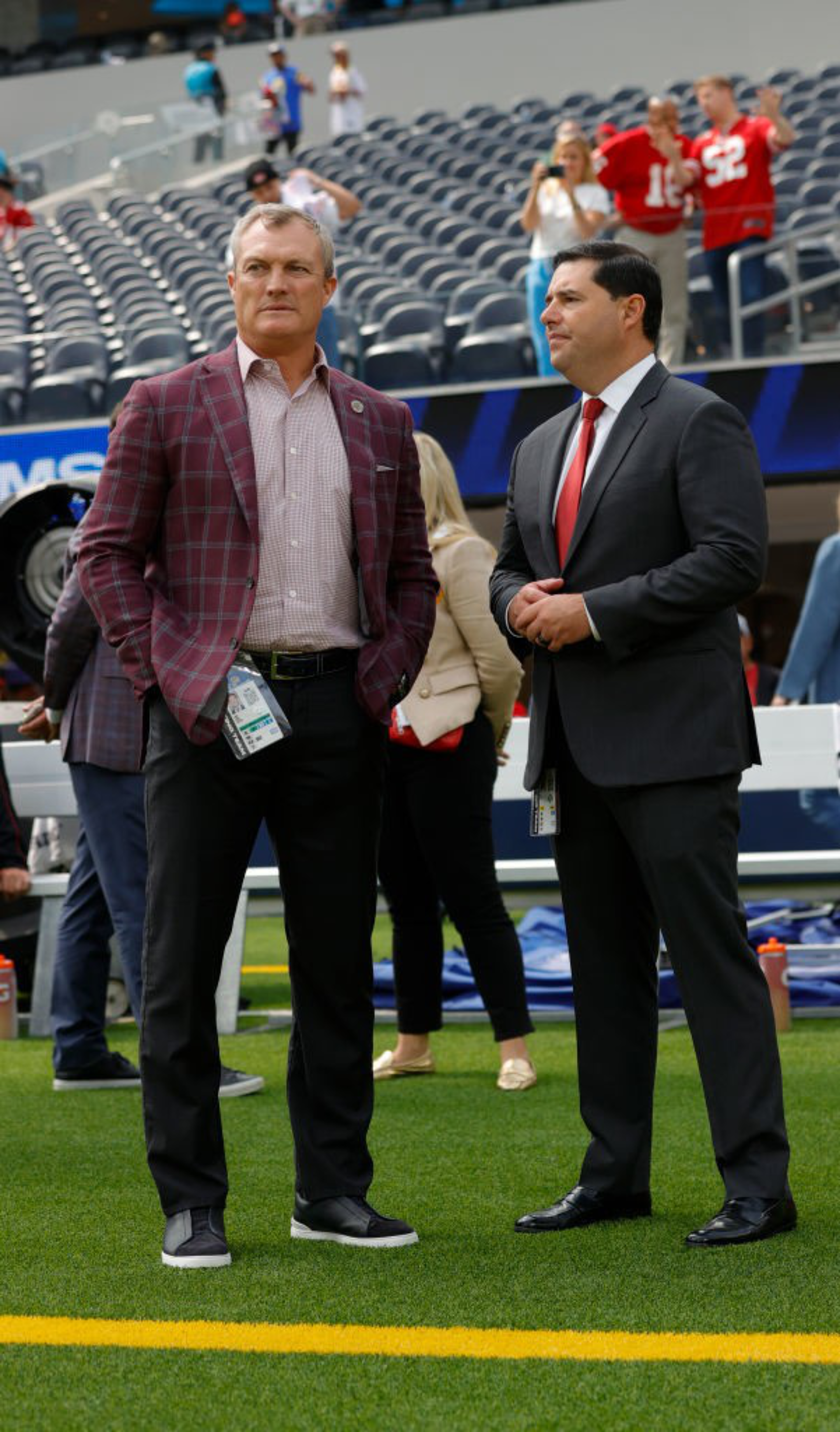 Two men in suits stand on a football field, with one wearing a plaid jacket and the other a red tie. People in the background occupy stadium seats.